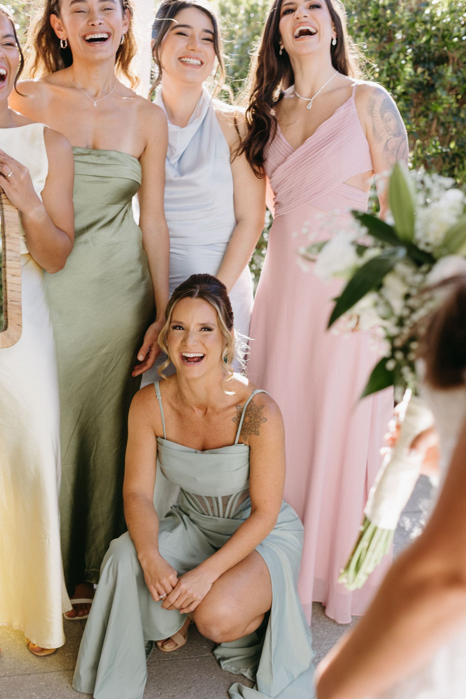 Bridesmaids in pastel green and pink dresses share candid laughing moments outdoors.