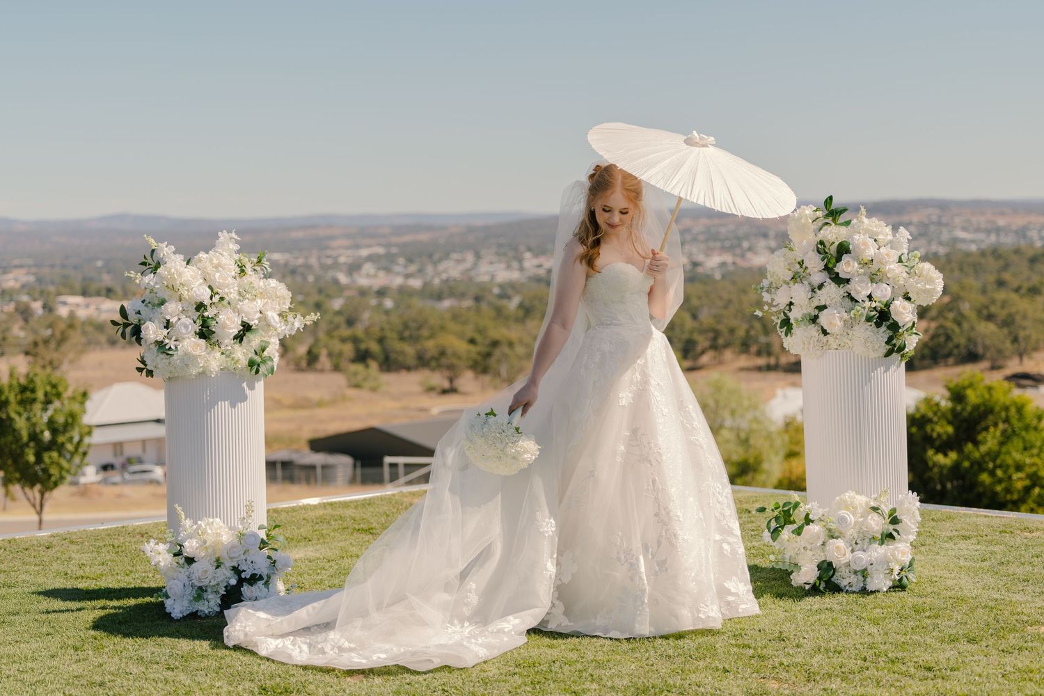 Bride in white ball gown holding parasol stands between floral arrangements at outdoor wedding ceremony.