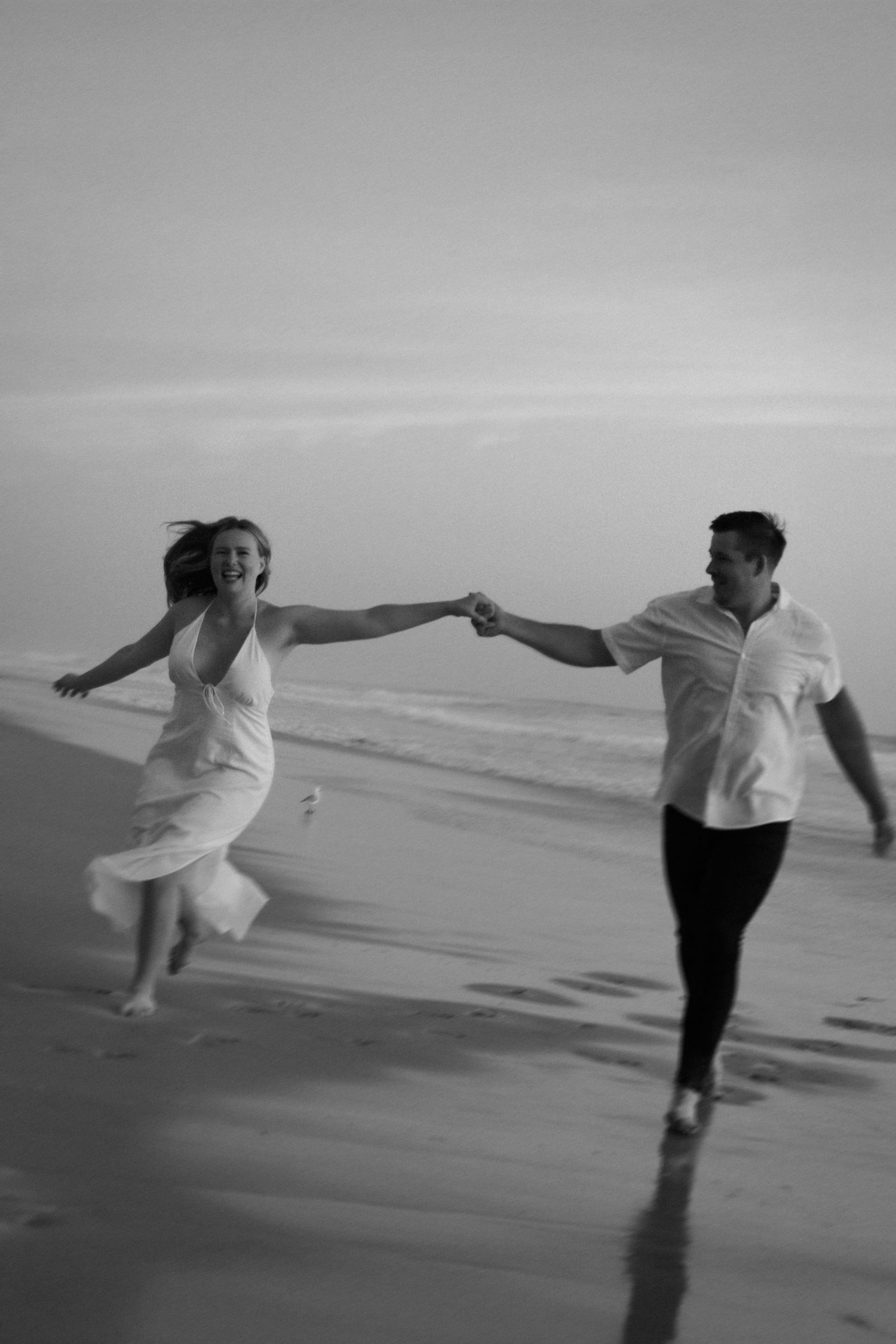 A couple in white clothing runs joyfully along a misty beach shore holding hands in black and white photography.