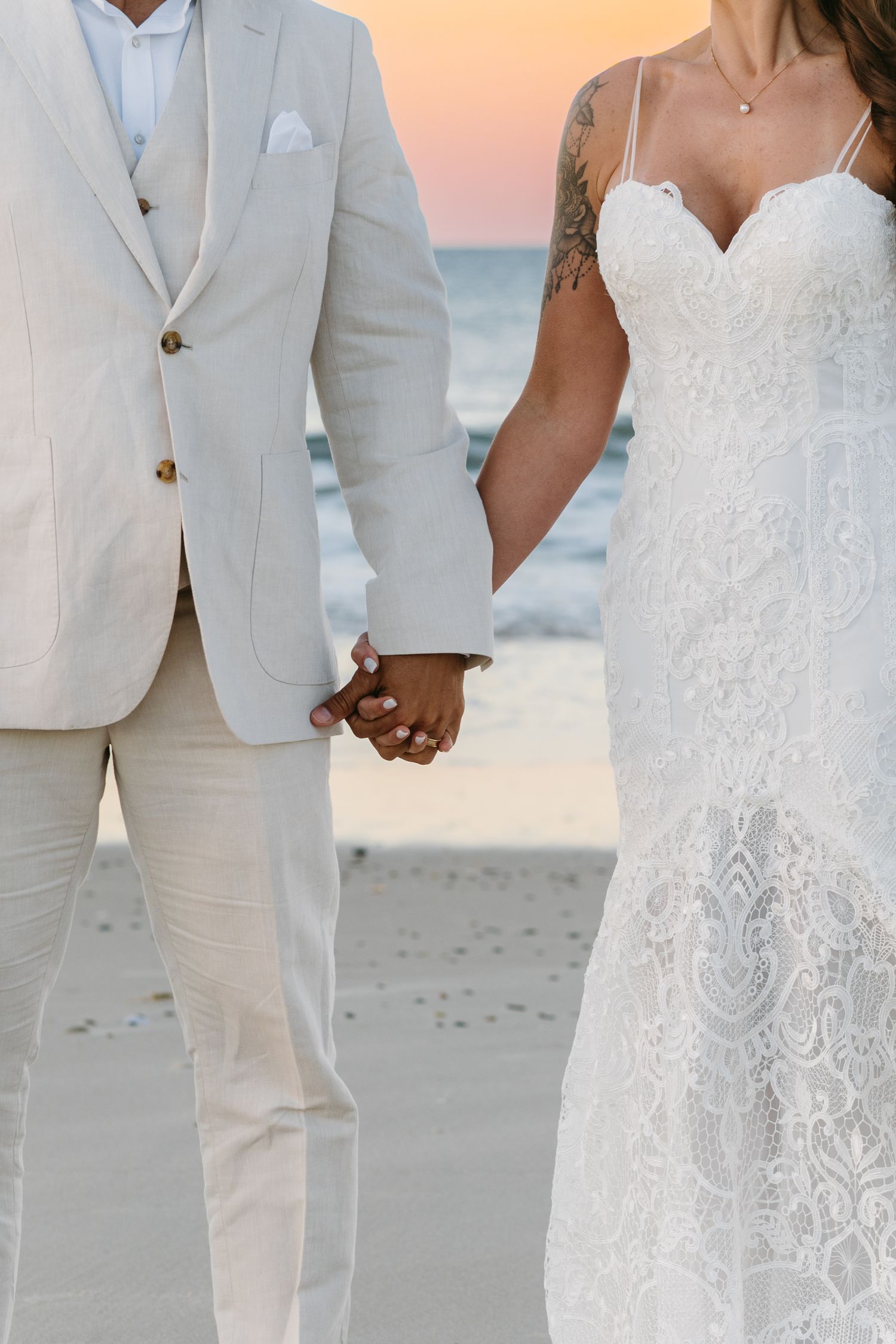 Couple in white wedding attire holding hands on a beach at sunset with romantic lighting.