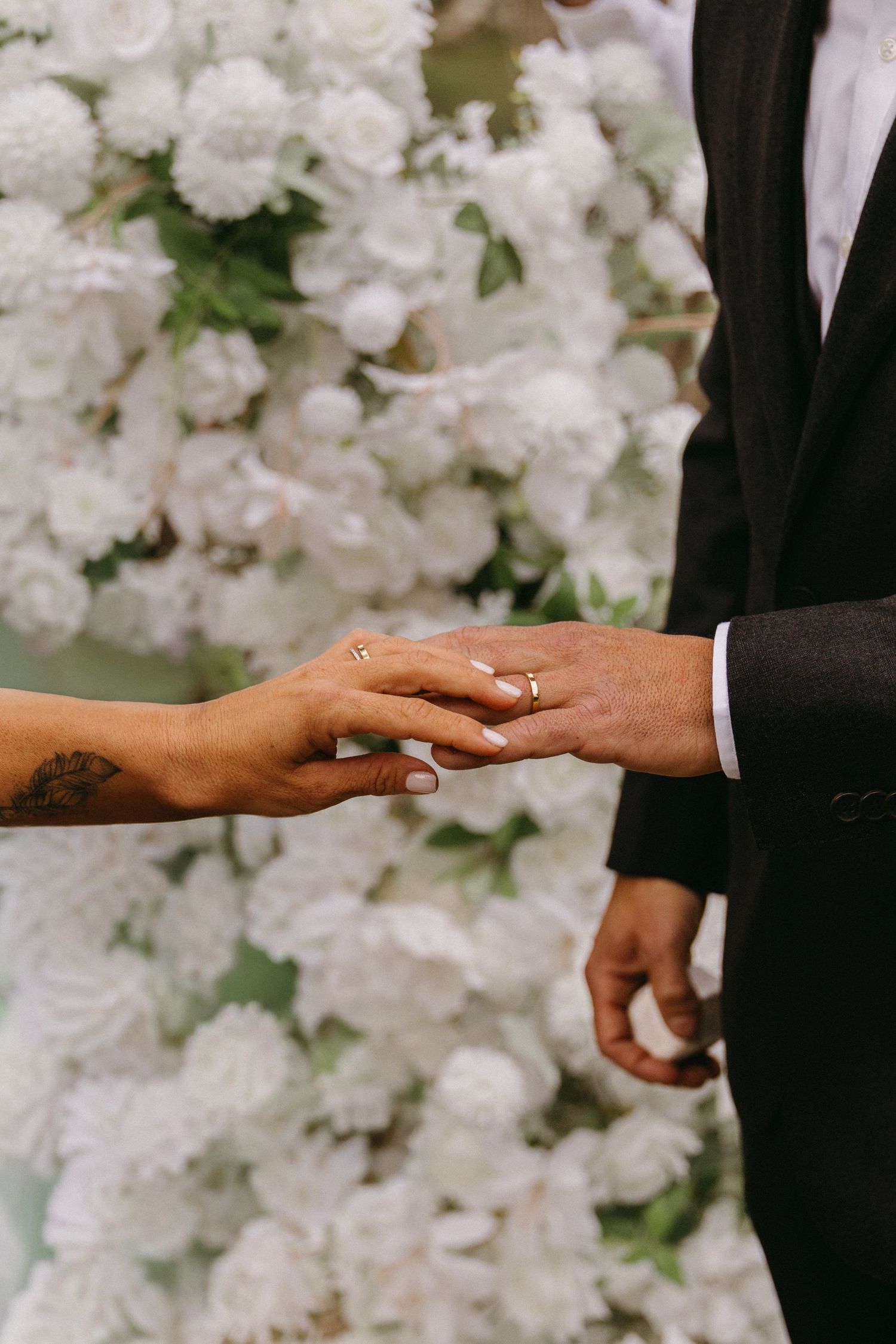 Two hands touch tenderly during a wedding ceremony against a backdrop of white floral arrangements.