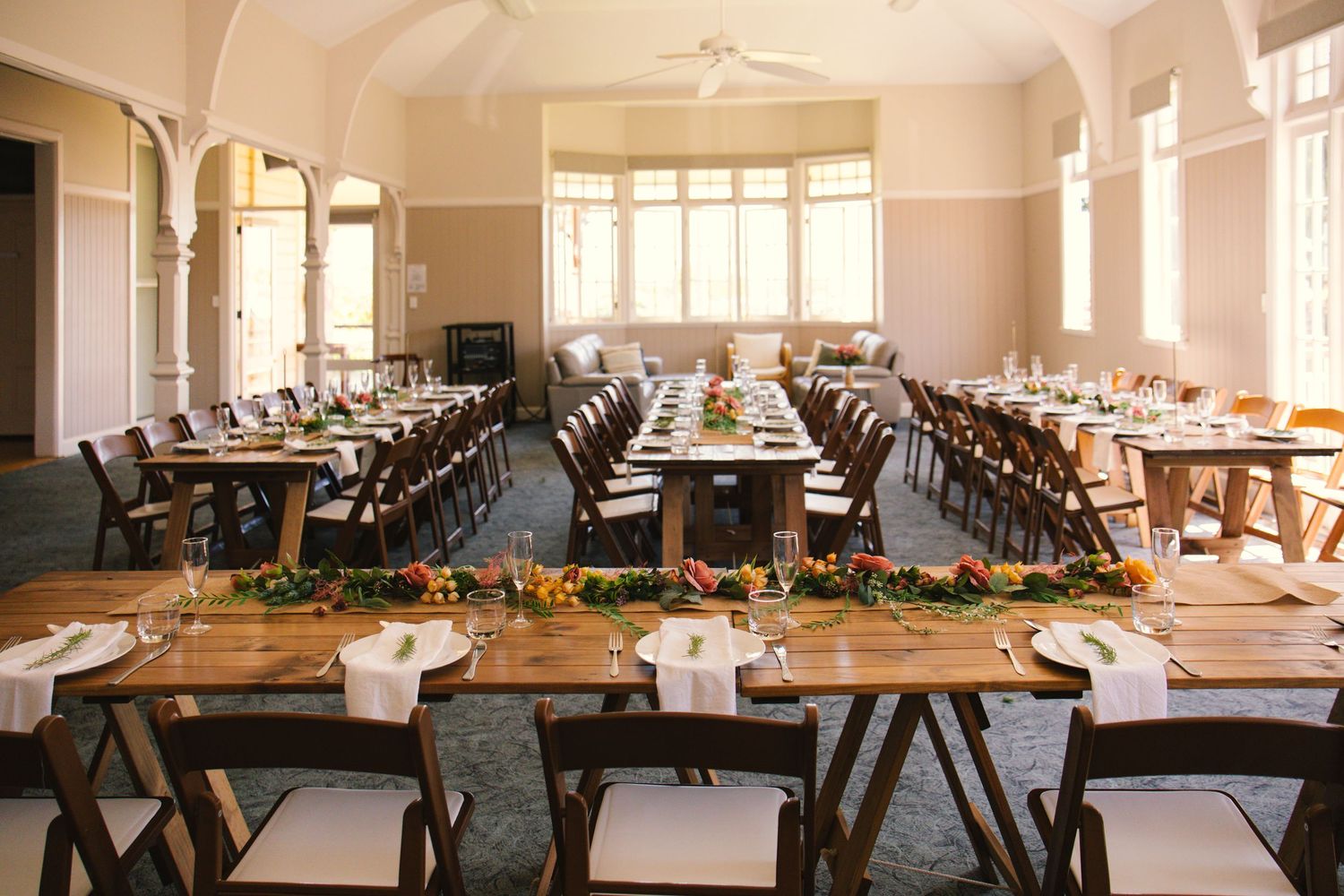 Long banquet tables set up in a bright, airy venue with large windows and white table settings.