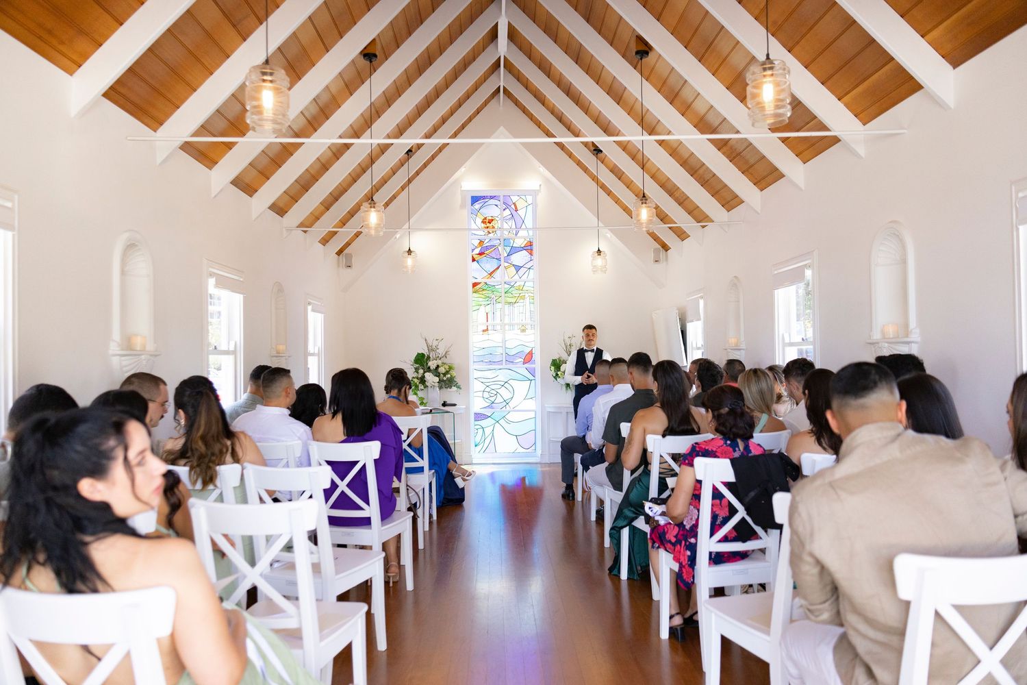 A bright white chapel interior with exposed wooden beams and rows of white chairs during a wedding ceremony.