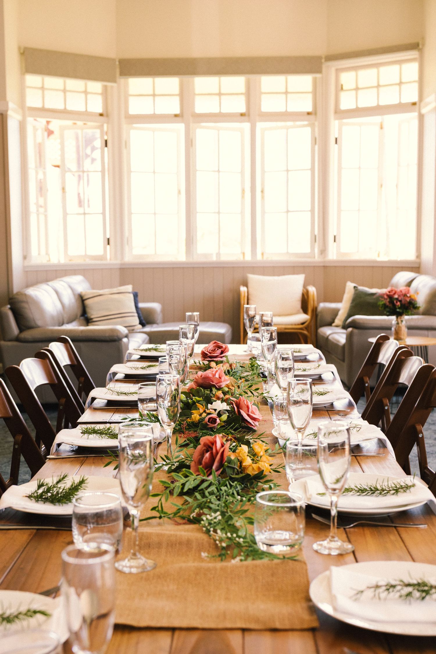 Elegant table setting with rustic wooden table, white plates, and natural greenery garland centerpiece in sunlit dining room.