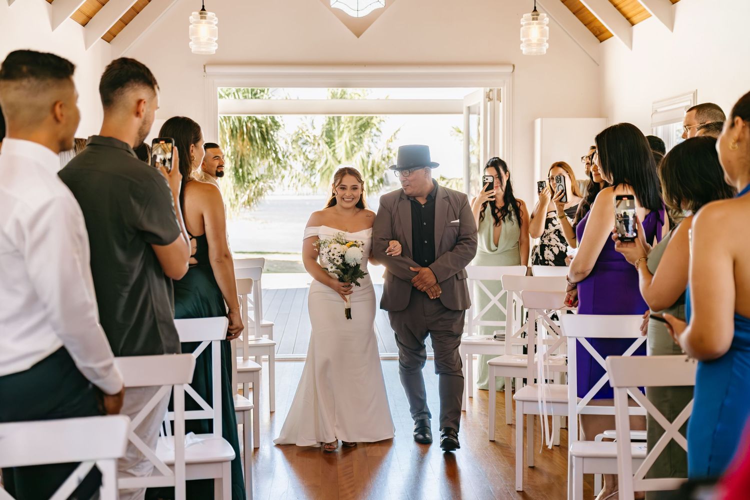 Wedding guests dance and celebrate together in a bright, sunlit reception venue with white decor and wooden beams.