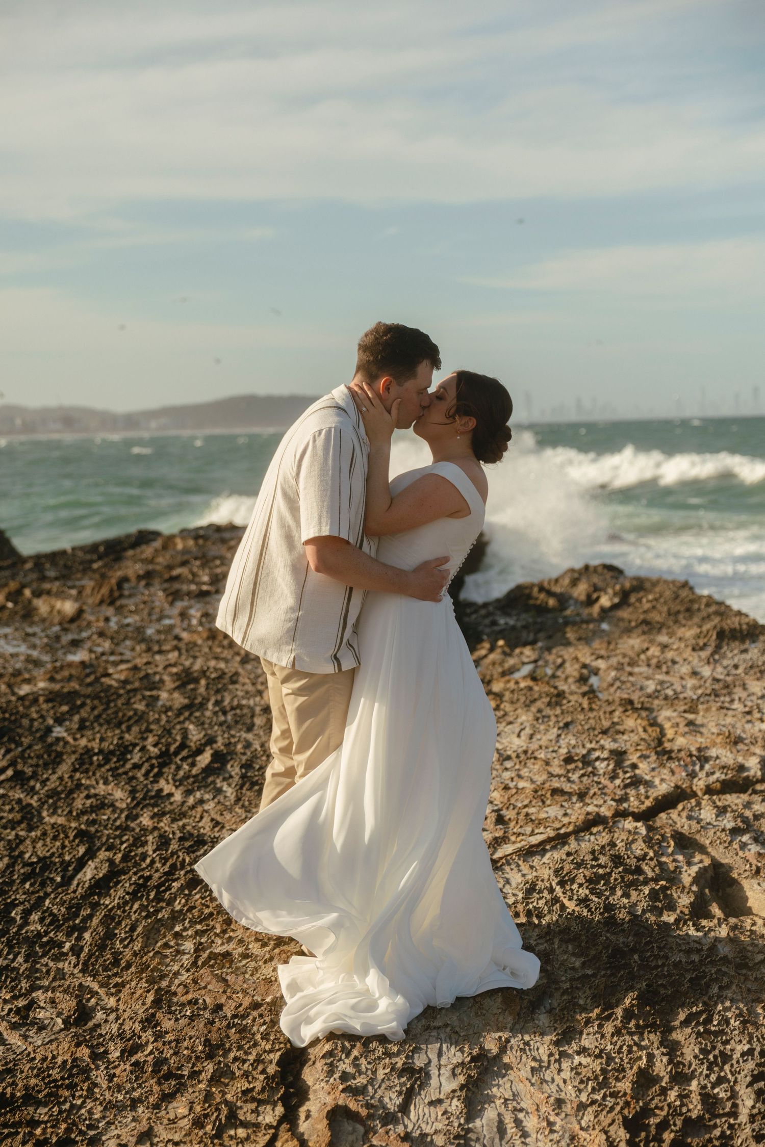 A couple in white attire shares an intimate moment on rocky coastal cliffs as ocean waves crash behind them.