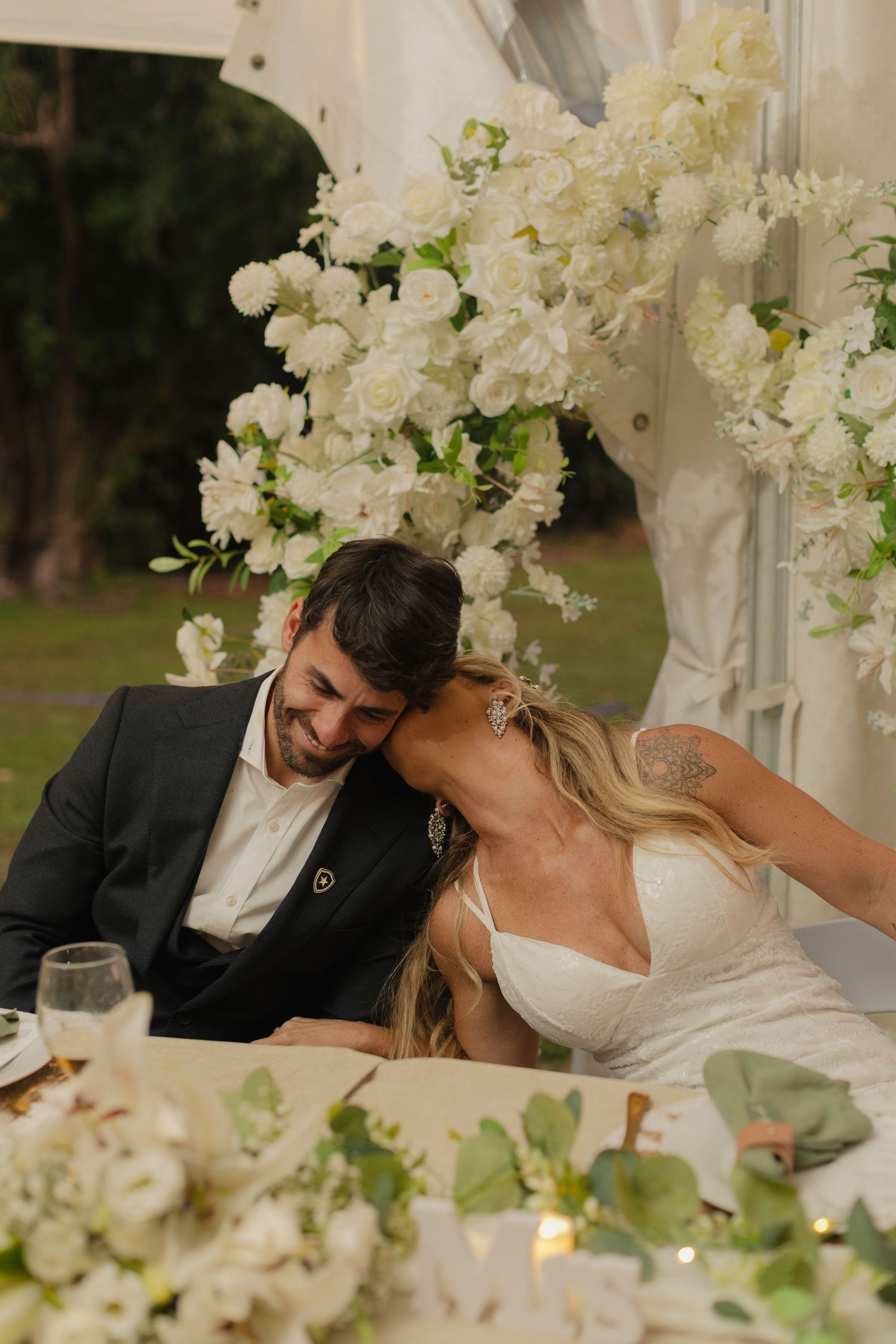 Newlyweds share intimate moments at a reception table decorated with white floral arrangements.