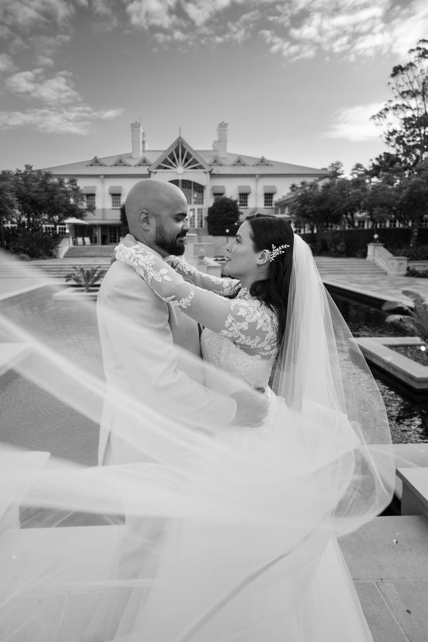 A romantic black and white wedding photo with a couple embracing in front of an elegant white mansion.