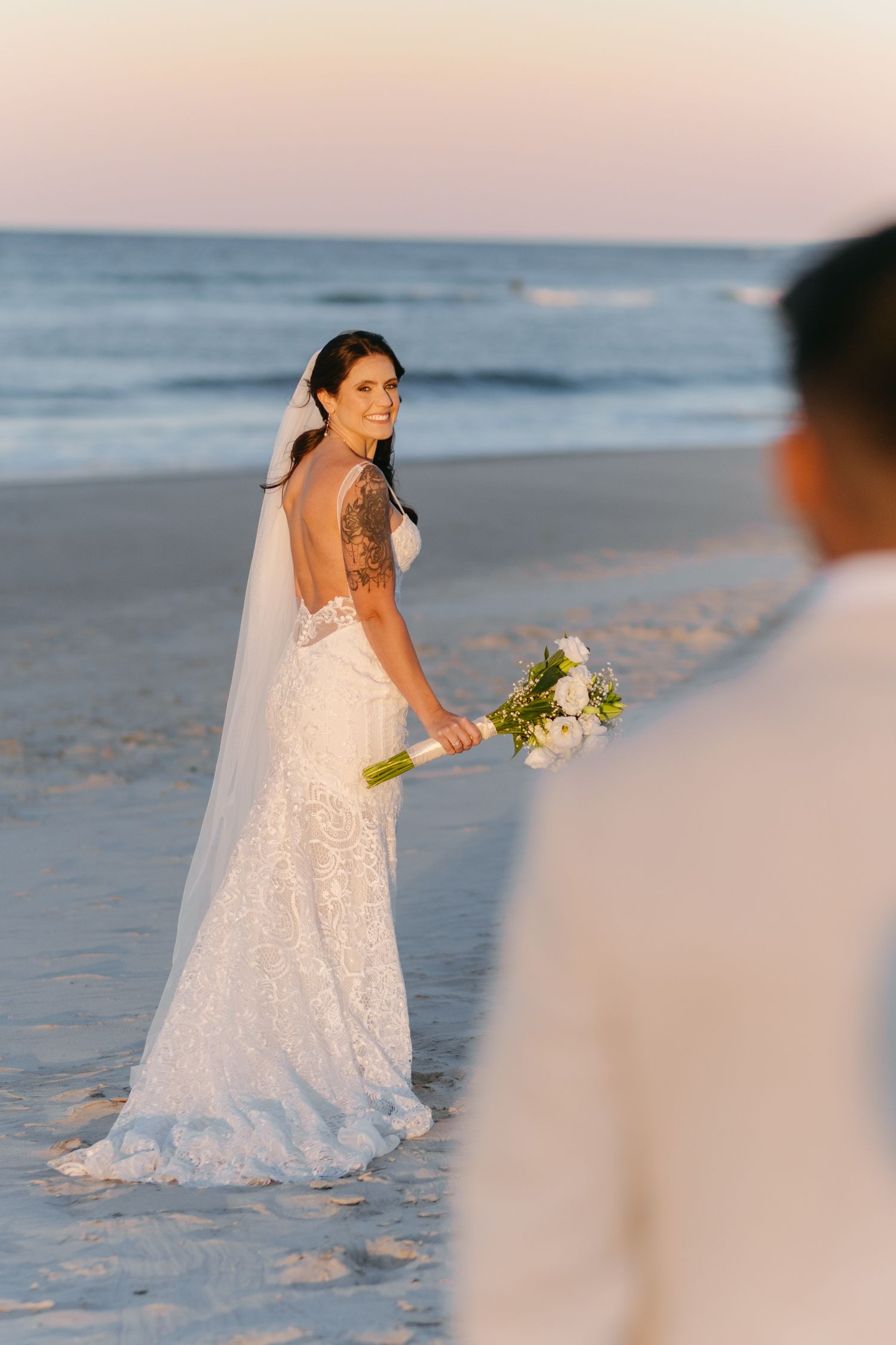 A bride in a lace wedding dress and veil holds a white bouquet while standing on a beach at sunset.