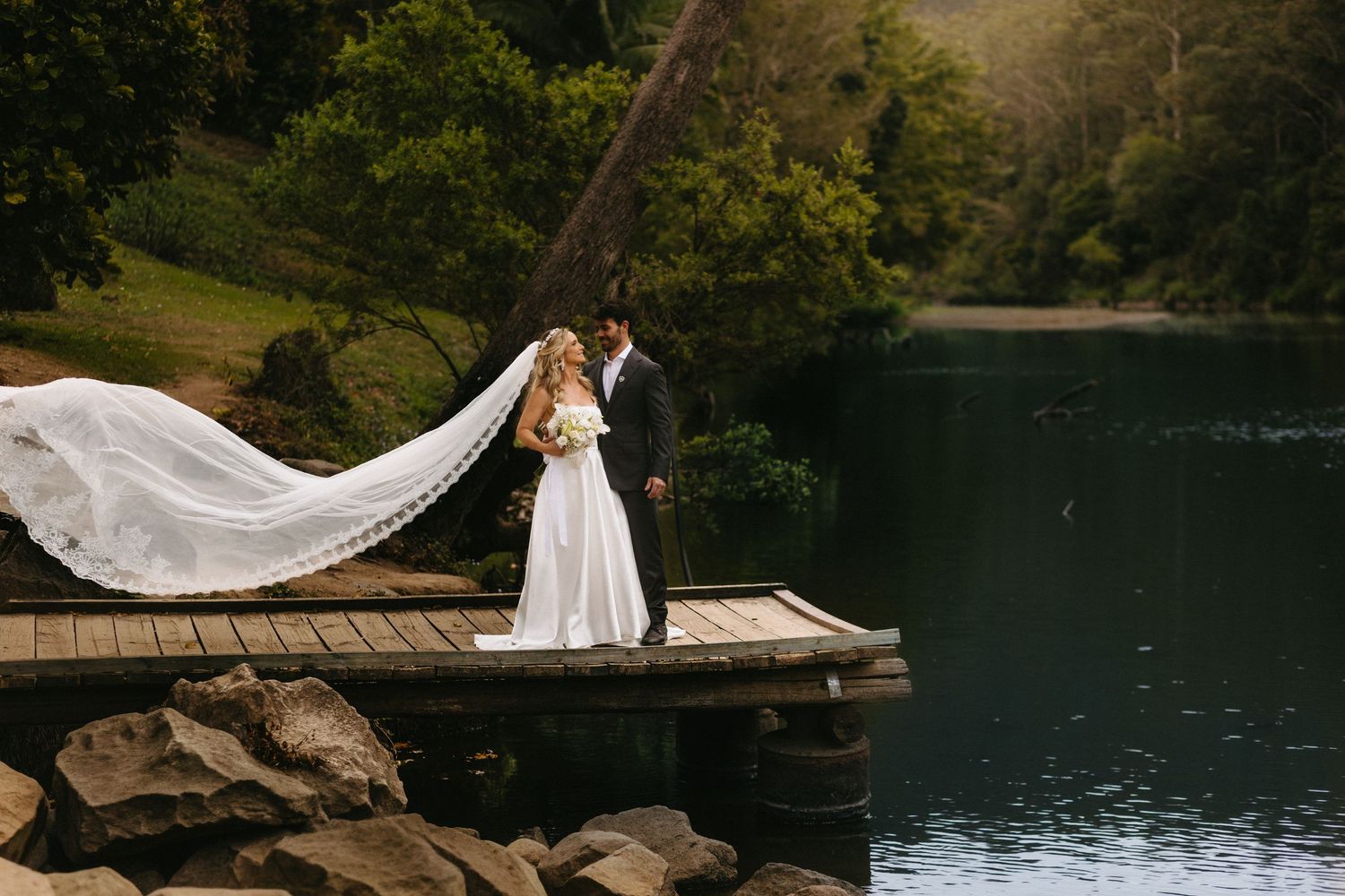 Romantic wedding couple poses on wooden dock by serene lake surrounded by lush greenery and mountains.