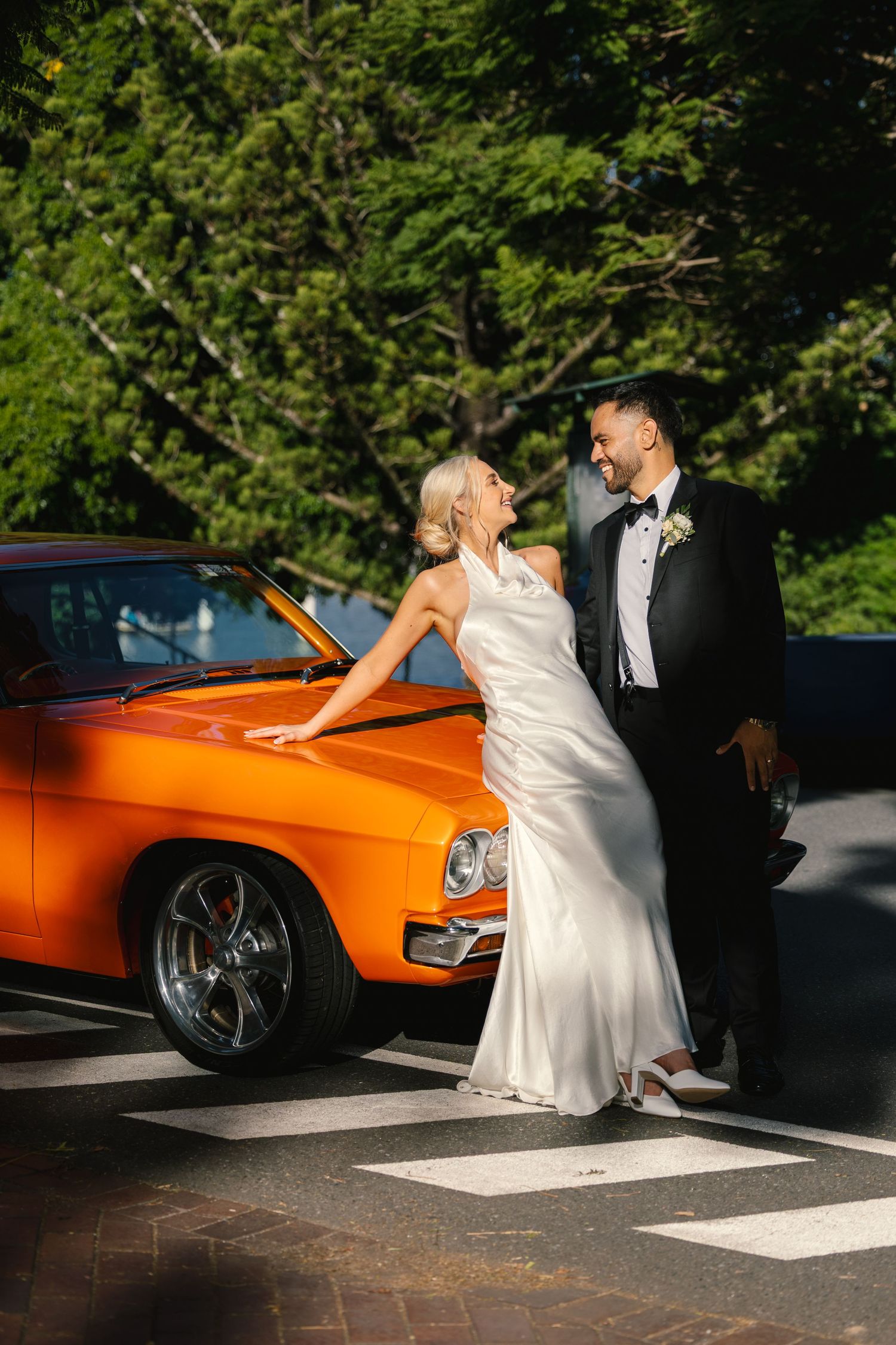 A couple in formal wedding attire pose beside a bright orange classic car in a sunlit parking lot.