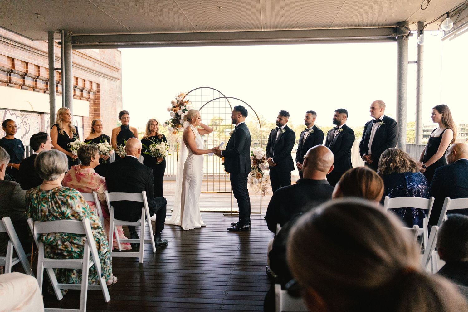 Wedding guests stand in rows during an outdoor ceremony under a covered patio area.