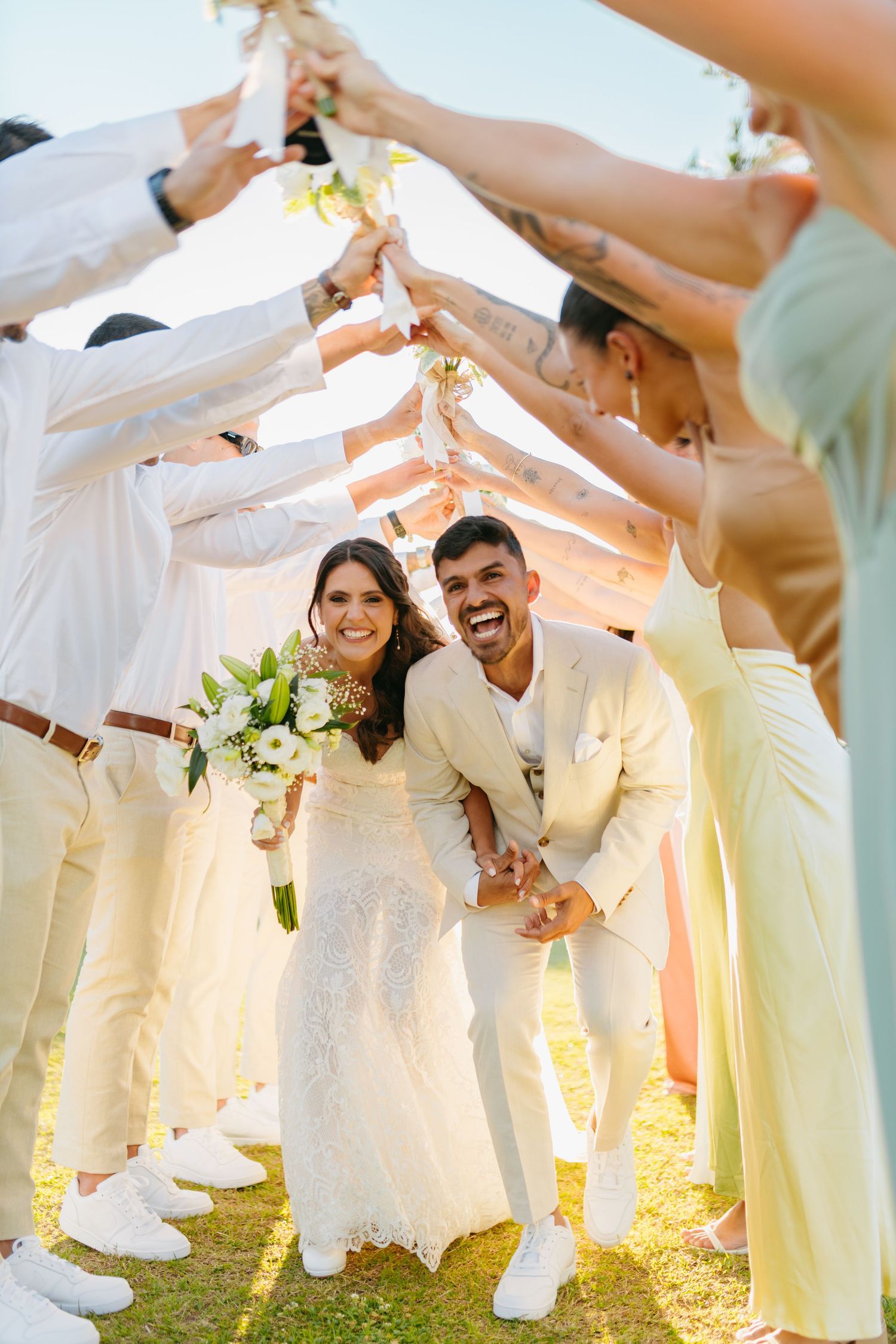 Wedding party in white and pastel yellow attire forms a tunnel for celebration under white fabric in golden sunlight.