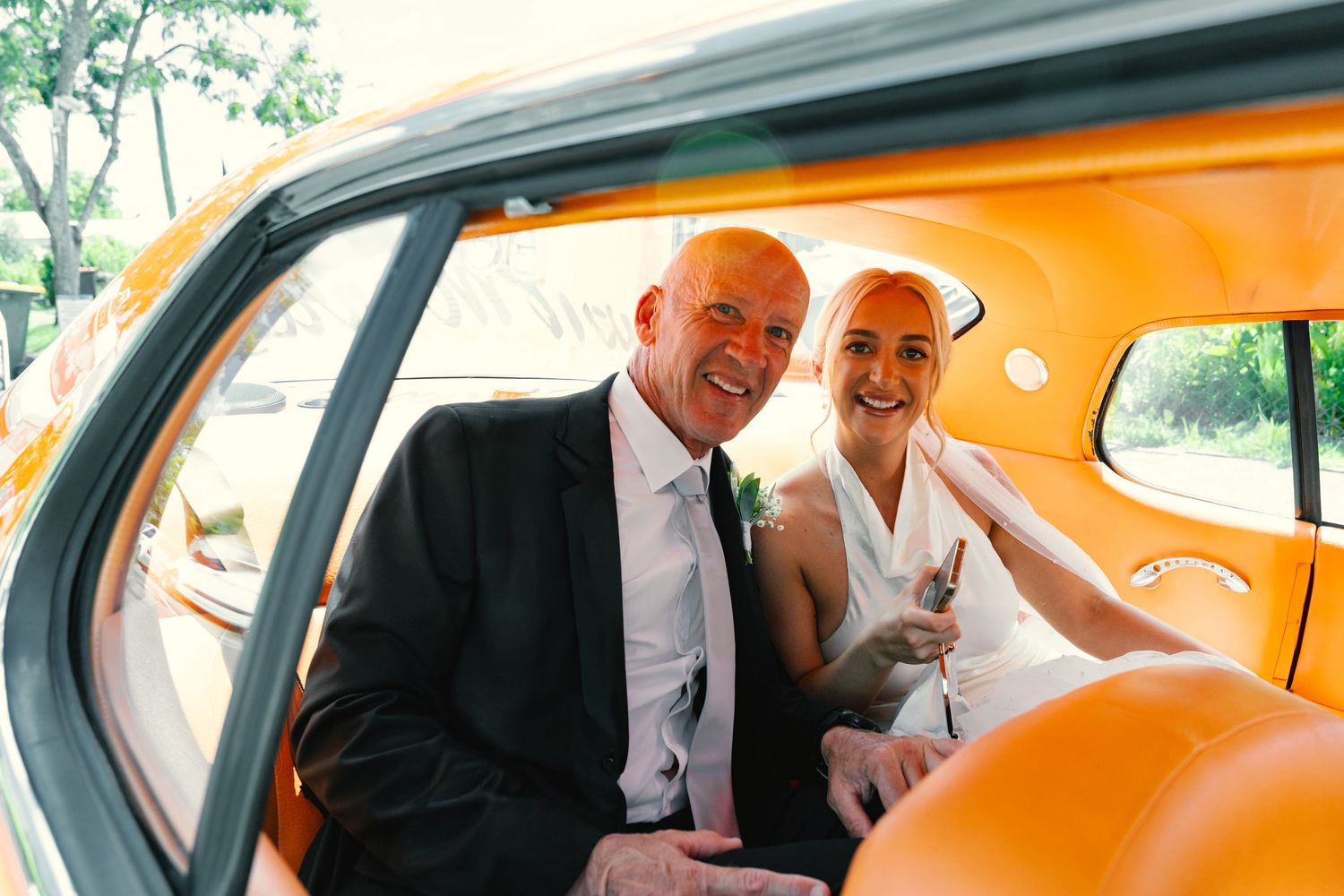 Wedding couple shares a joyful moment in the back seat of a vintage orange car.