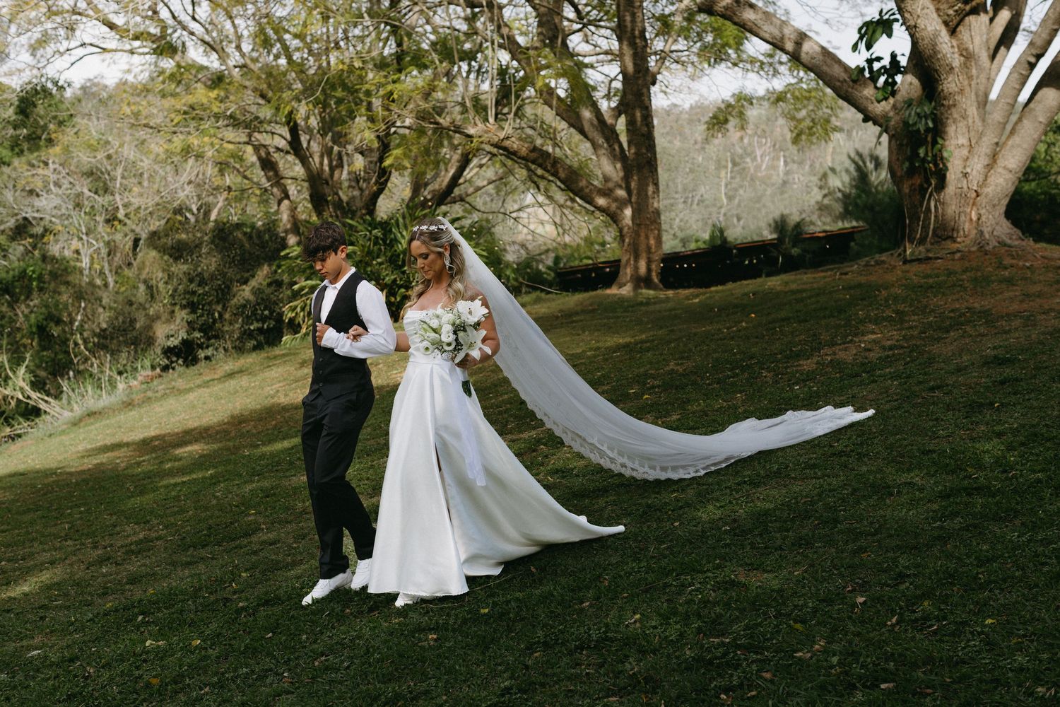 Bride's flowing veil catches the breeze during outdoor wedding photos under mature trees.
