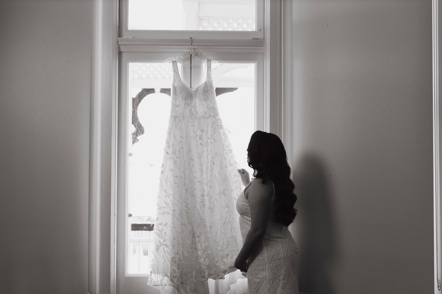 Black and white silhouette photo showing wedding dress preparations through window light.