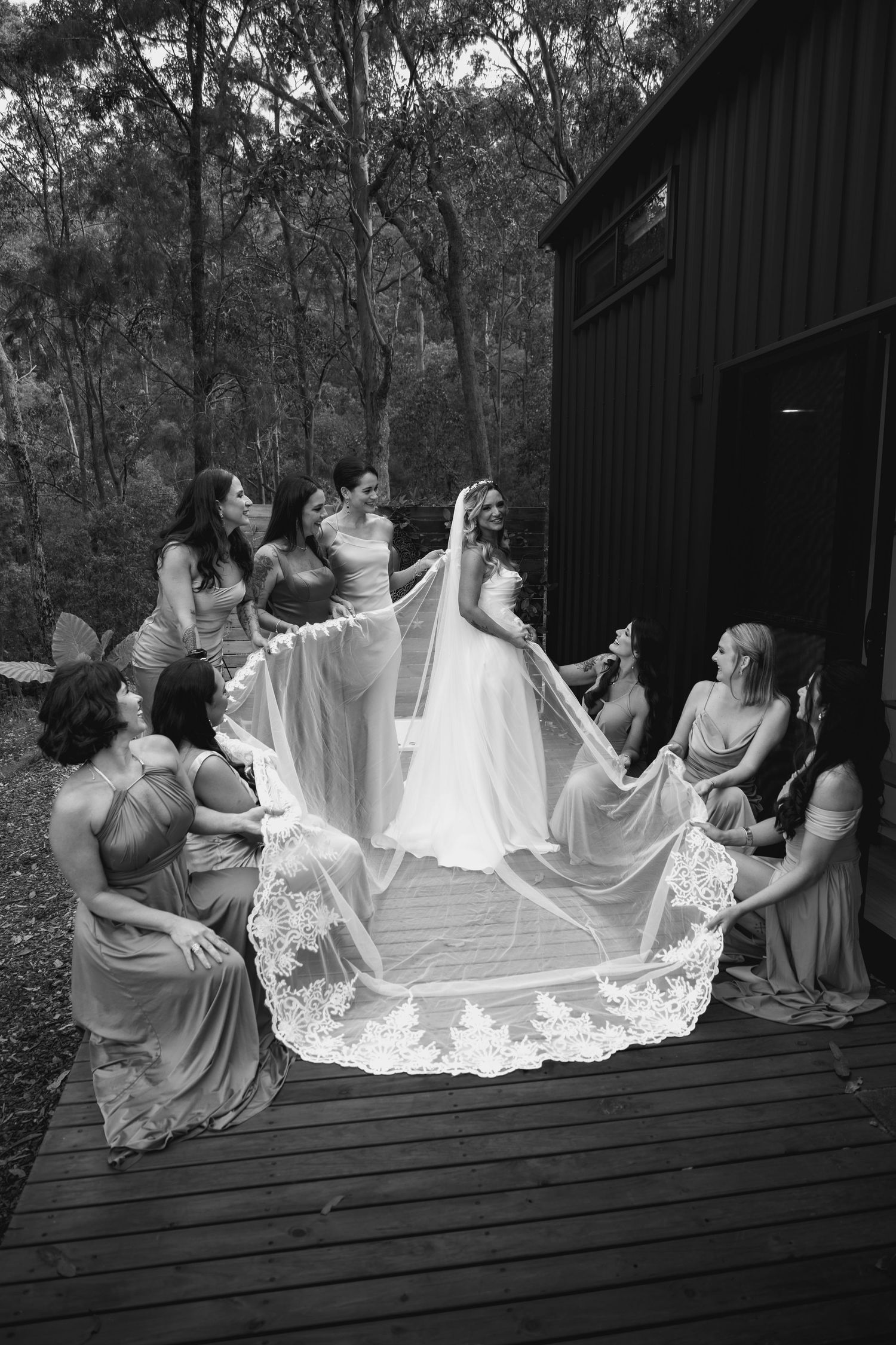 Wedding party arranges the bride's lace veil on a wooden deck in black and white photography.