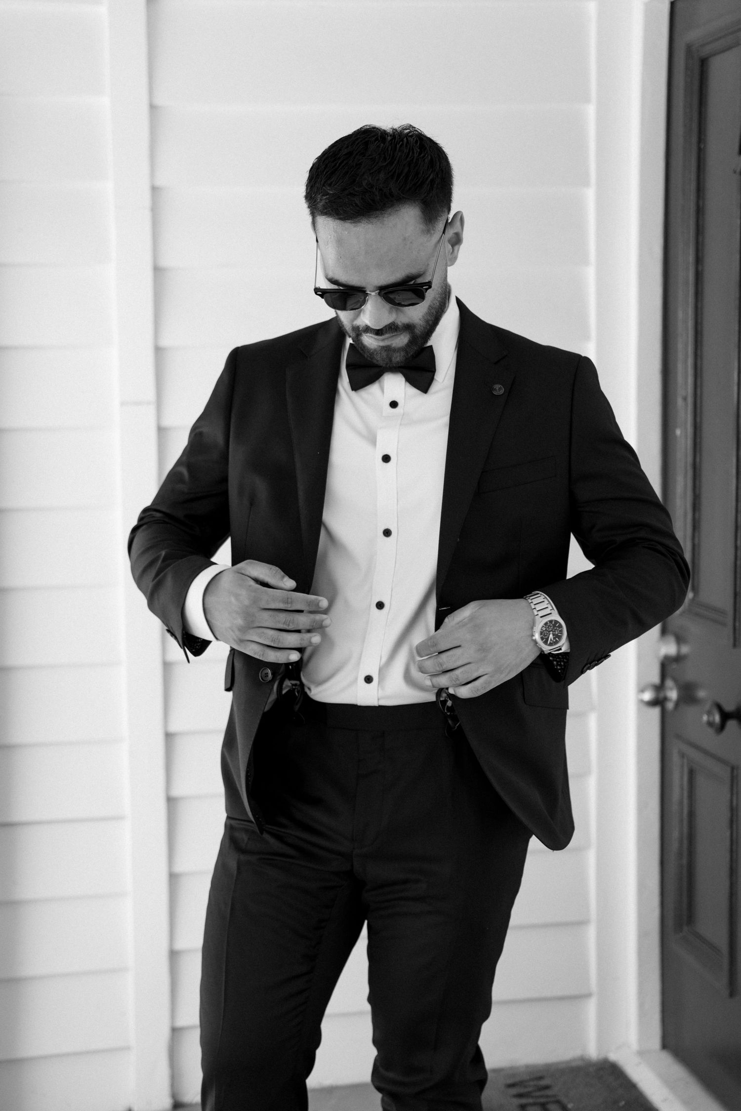 Groom in classic black tuxedo and bow tie adjusts his jacket in black and white photo.