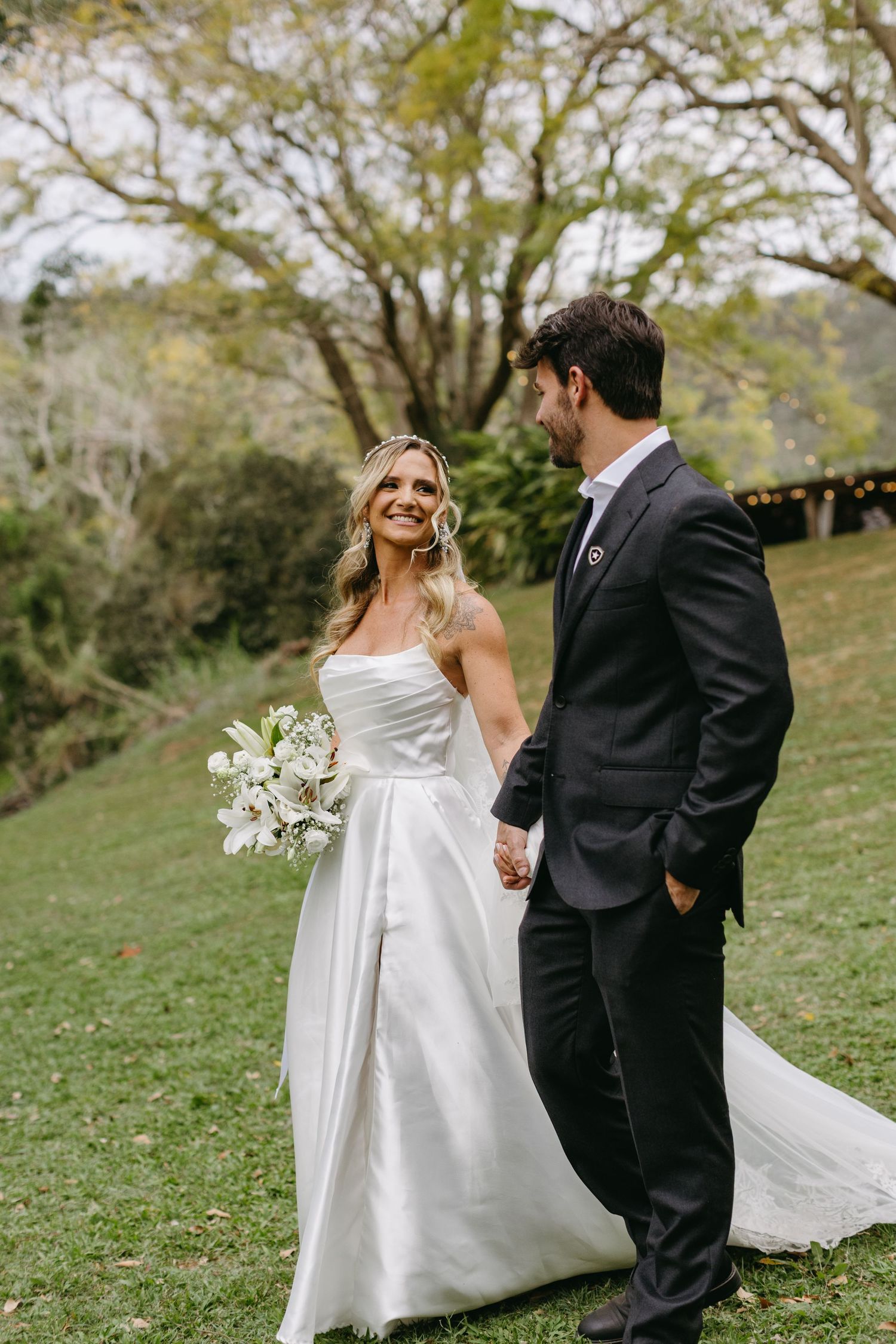 A bride in a flowing white dress holds a bouquet while walking with her groom on a grassy hillside.