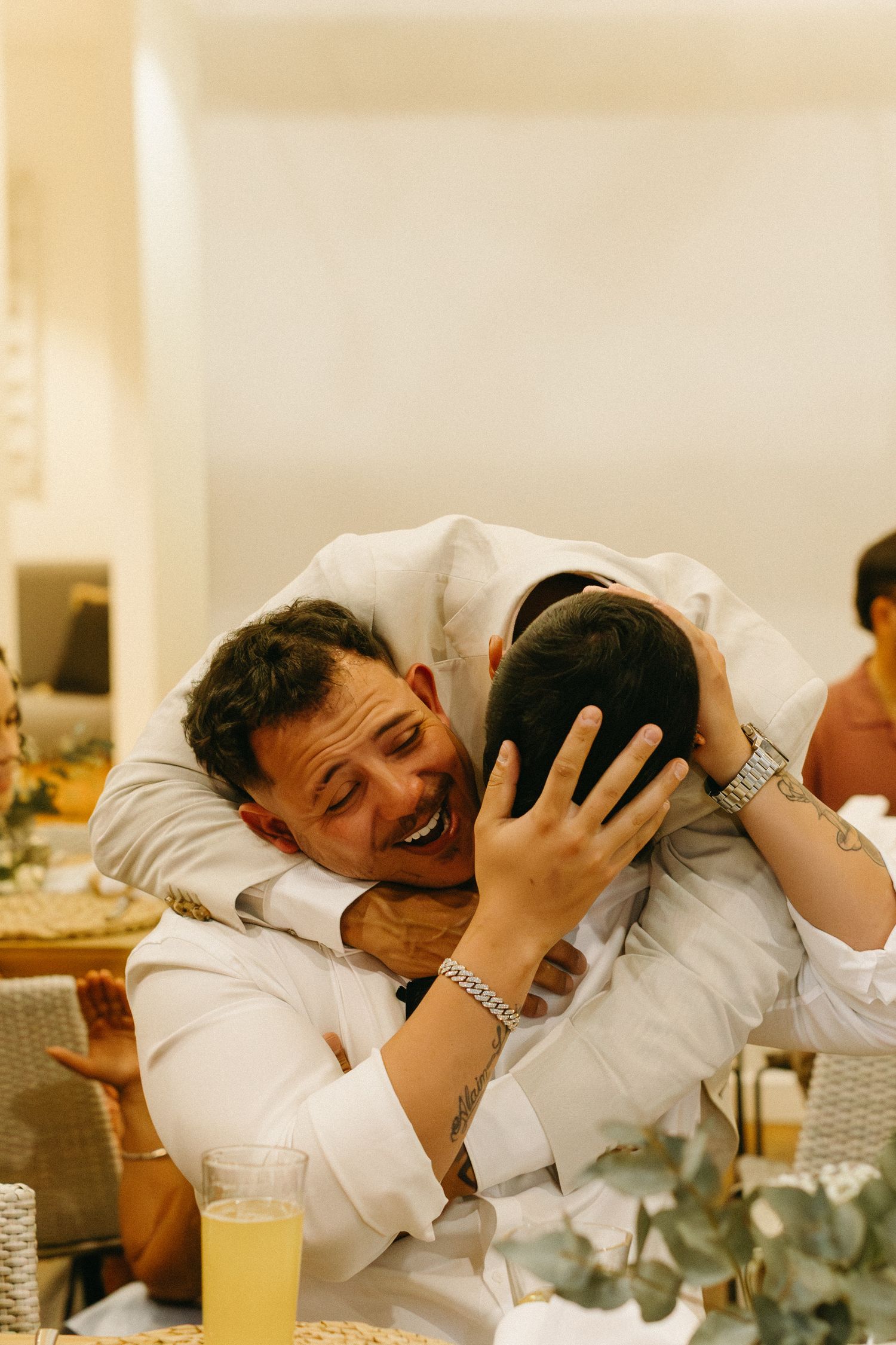 Two people in white clothing share an emotional embrace at what appears to be a wedding reception.