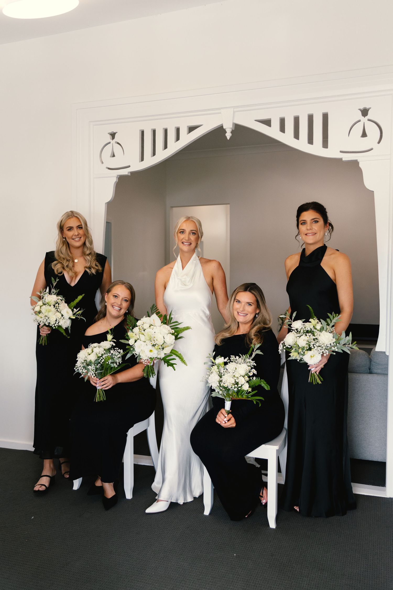 Wedding party poses together with bridesmaids in black dresses holding white floral bouquets.