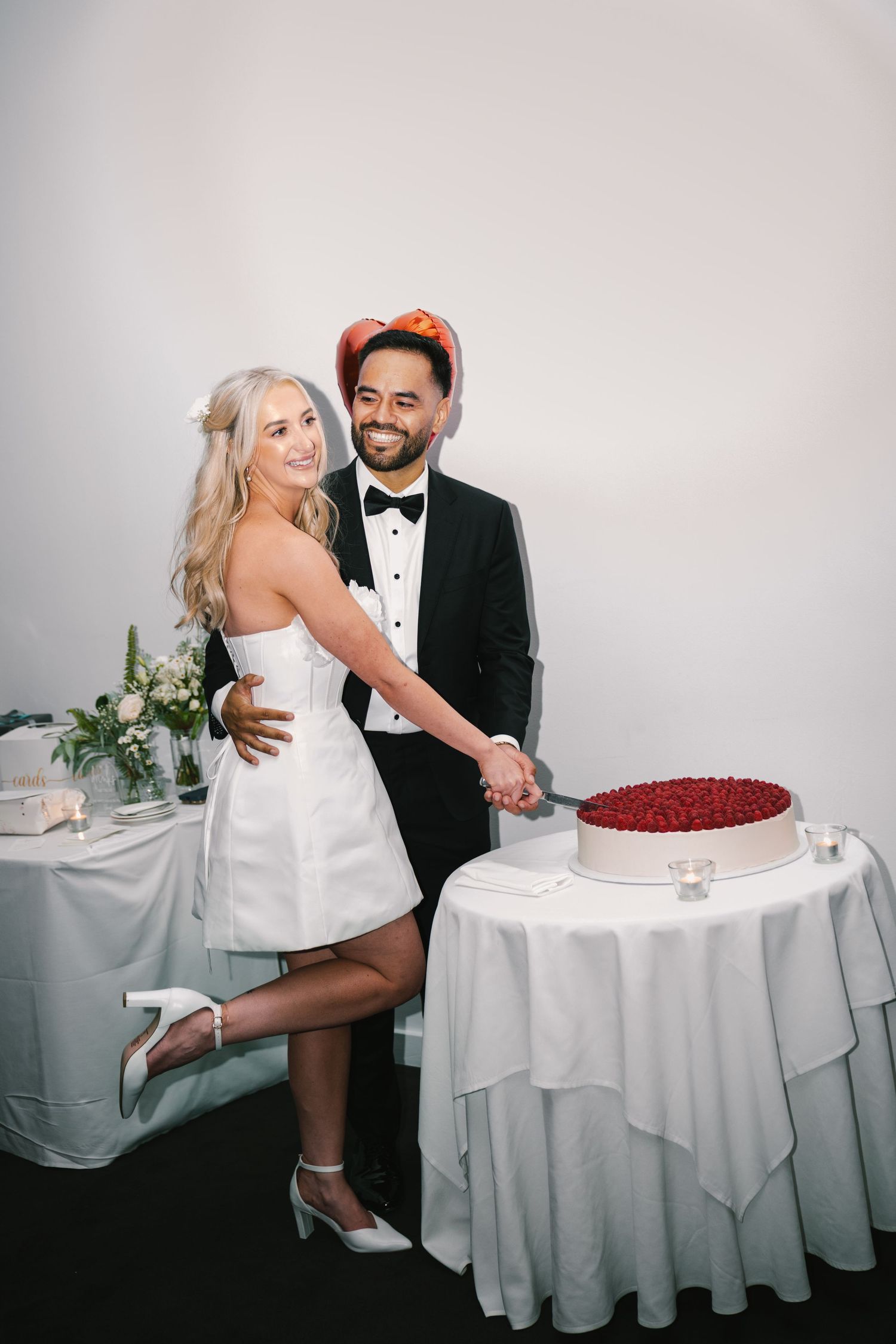 A couple poses together at their wedding reception next to a red dessert cake on a round table with white linens.