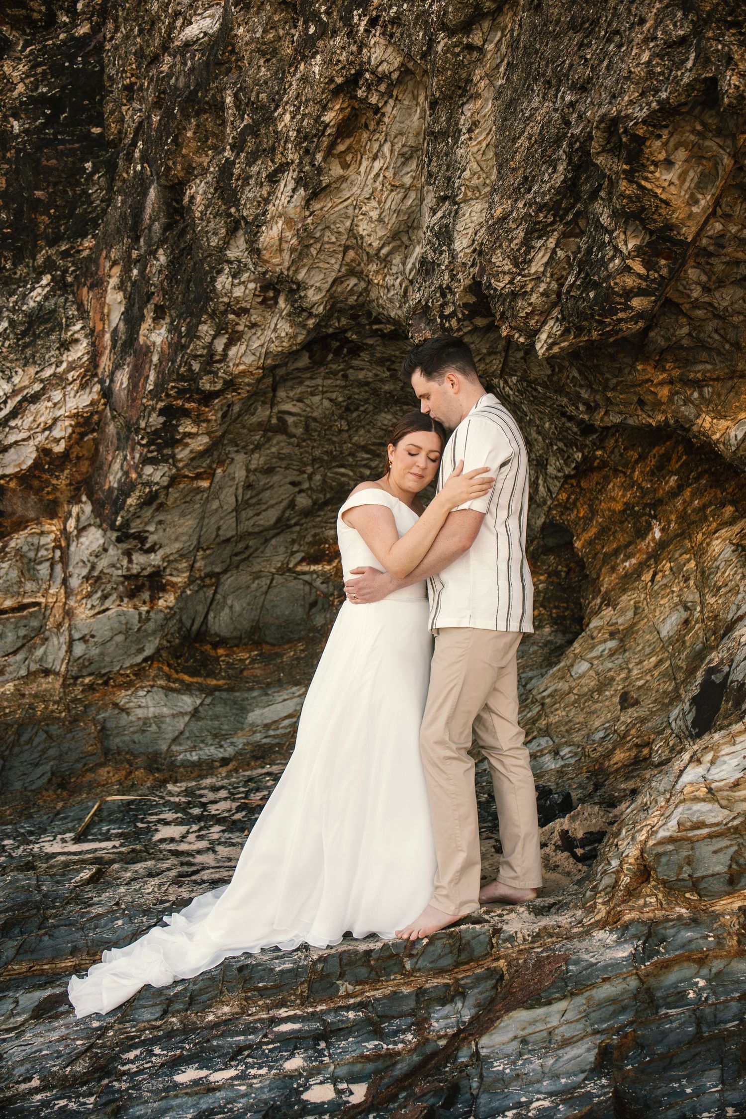 A romantic photoshoot under a rocky coastal cave with a couple in wedding attire posing together.