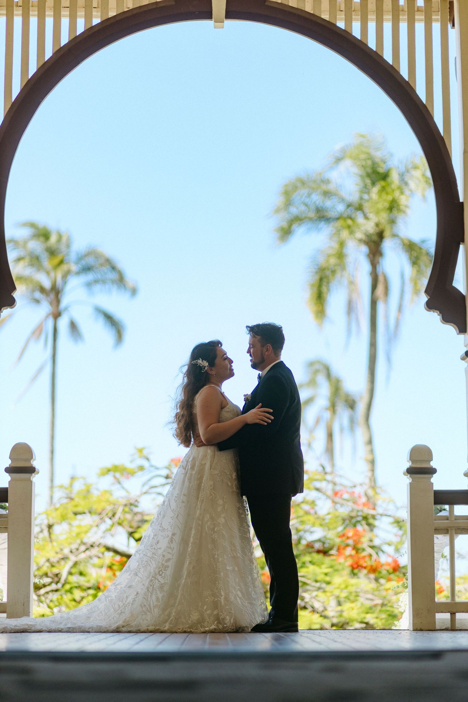 A bride and groom share romantic moments under an arched gazebo with palm trees and tropical flowers in the background.