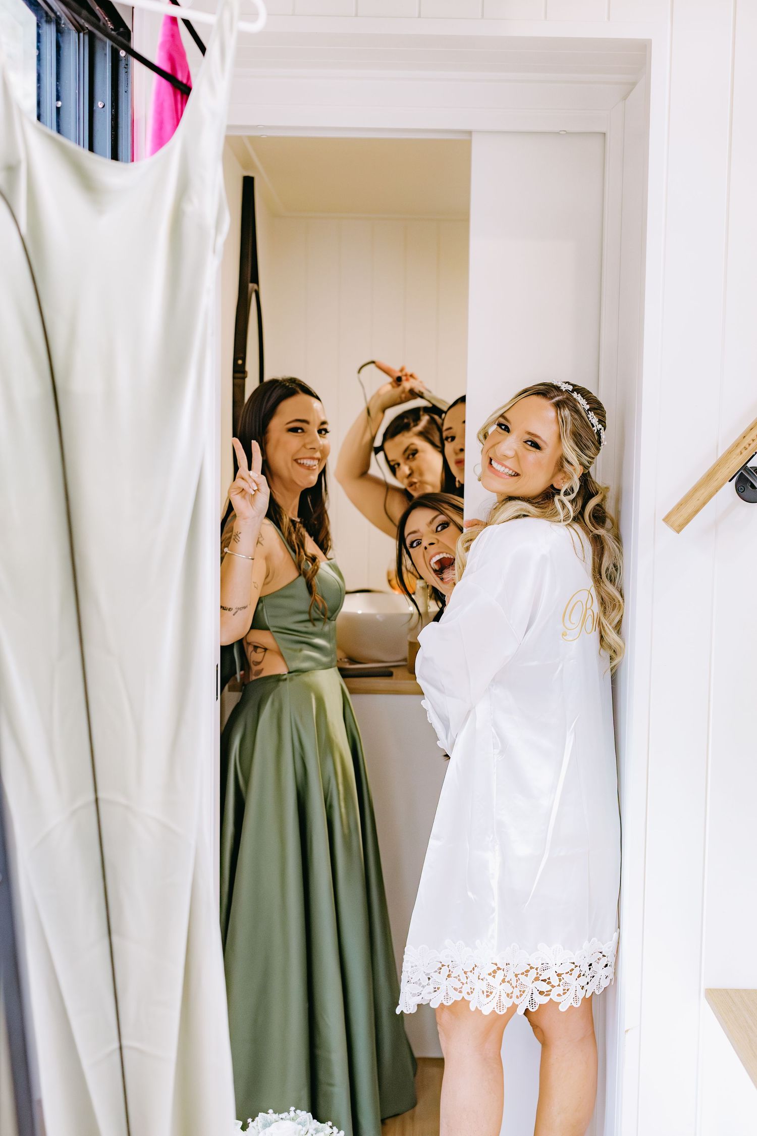 Wedding party getting ready in a bright white dressing room with sage green bridesmaid dresses visible.