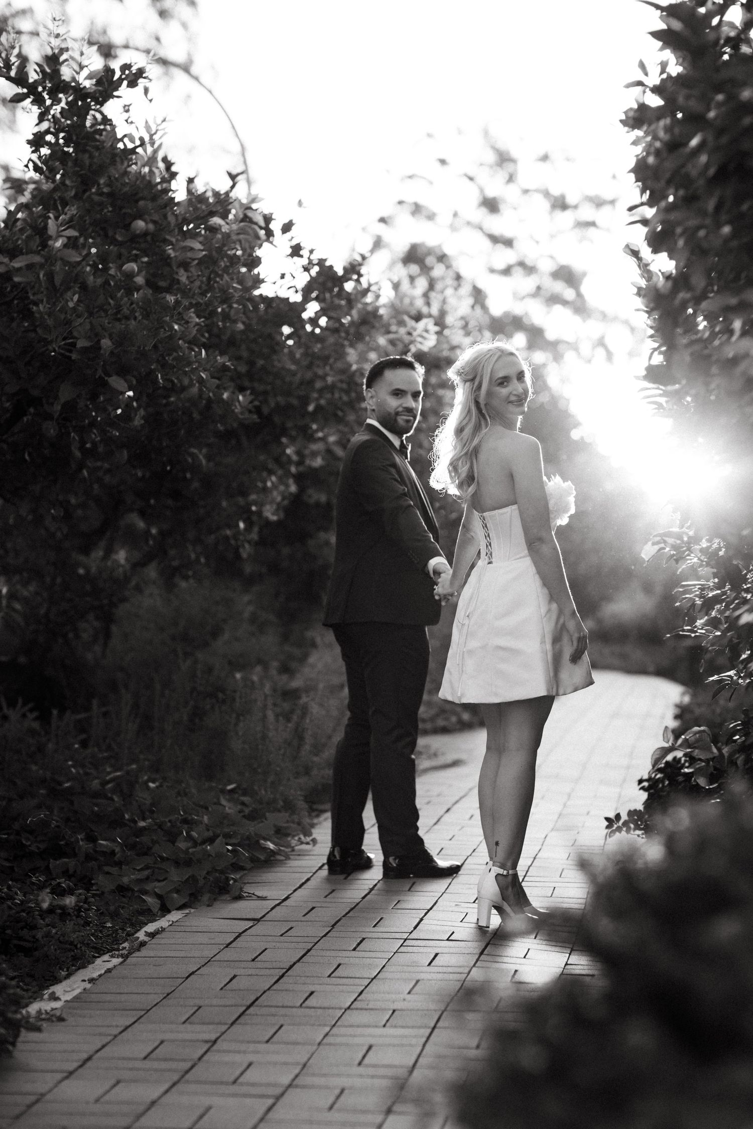 A wedding couple walks along a stone path in a garden with dramatic backlighting creating a romantic silhouette.