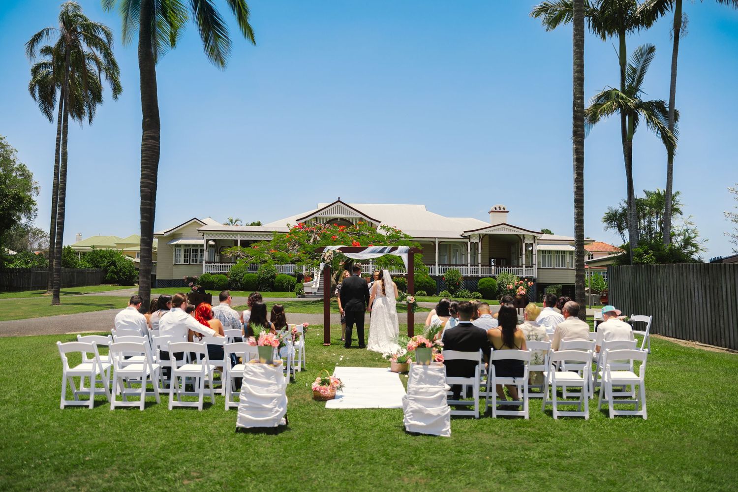 Outdoor wedding ceremony setup with white chairs arranged on a lawn under palm trees.