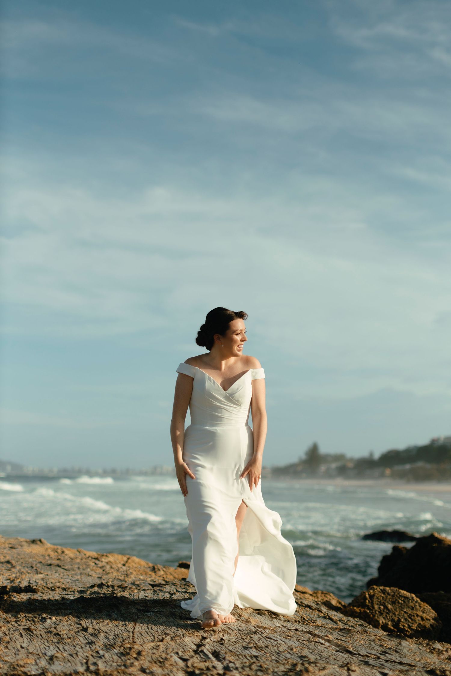 Person in flowing white dress sits gracefully on rocky coastal shoreline as waves crash behind them on cloudy day.
