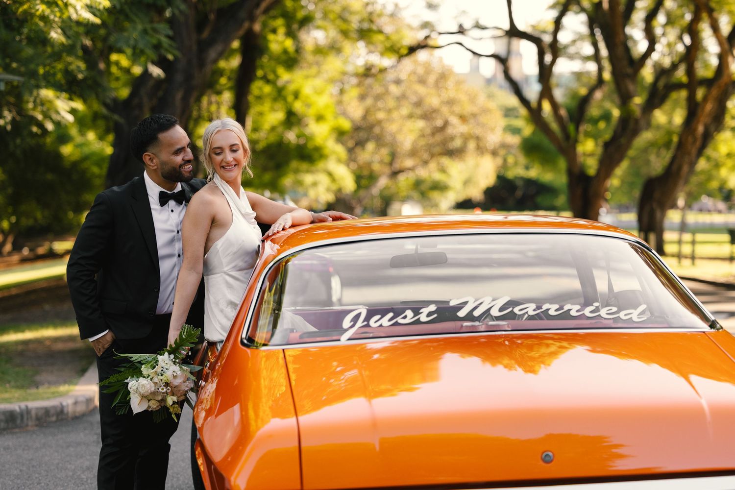 A classic orange car with 'Just Married' text on the window and a couple posing beside it in a park setting.