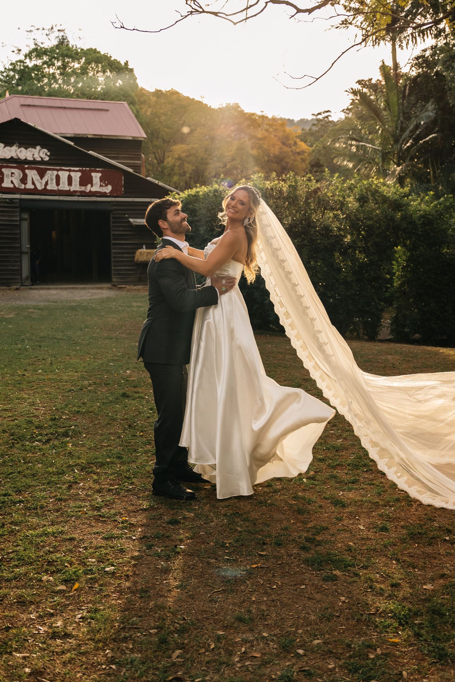 Couple dances at sunset near rustic barn with bride's veil flowing dramatically.