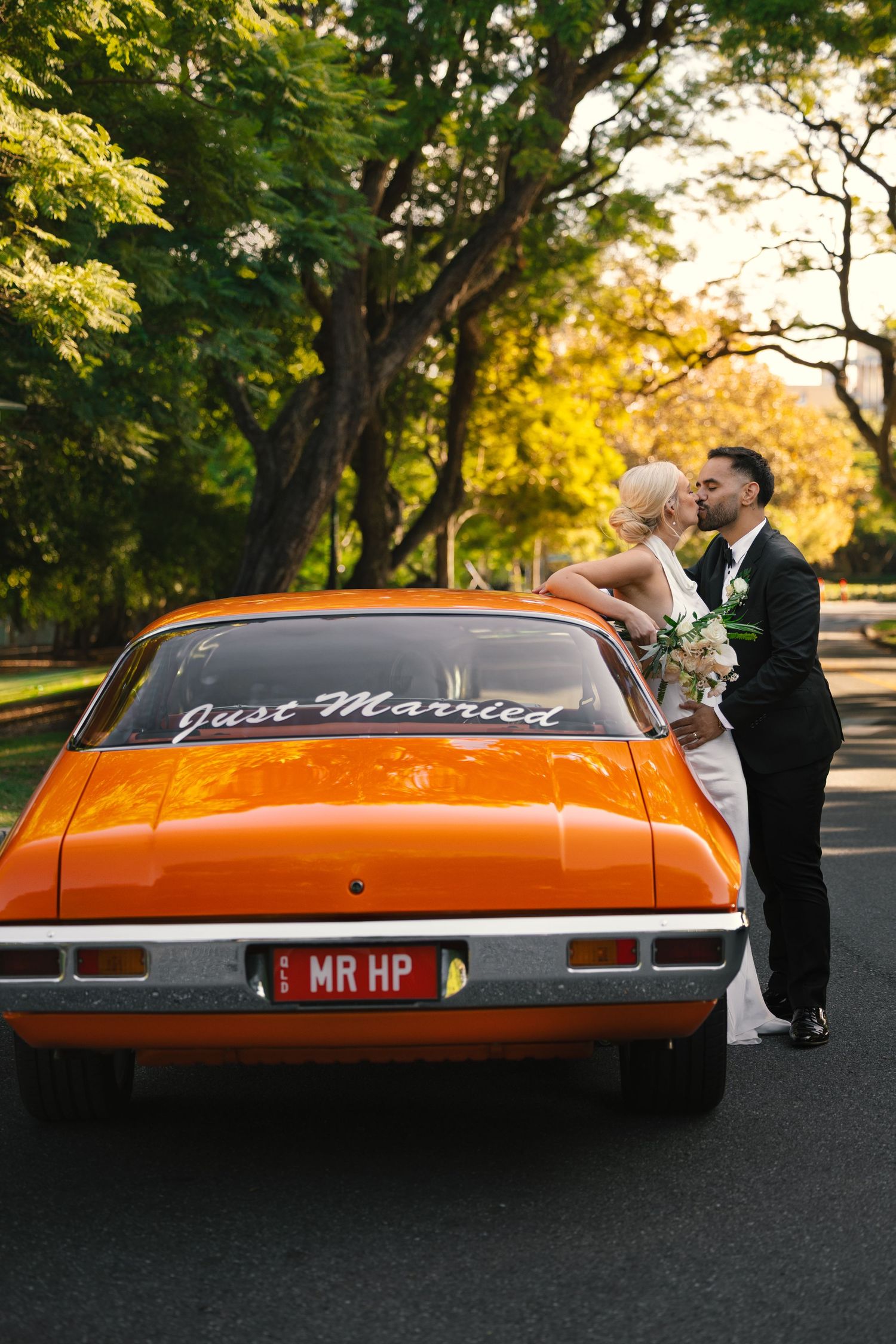 A classic orange Gran Torino car serves as a backdrop for wedding photos at sunset.