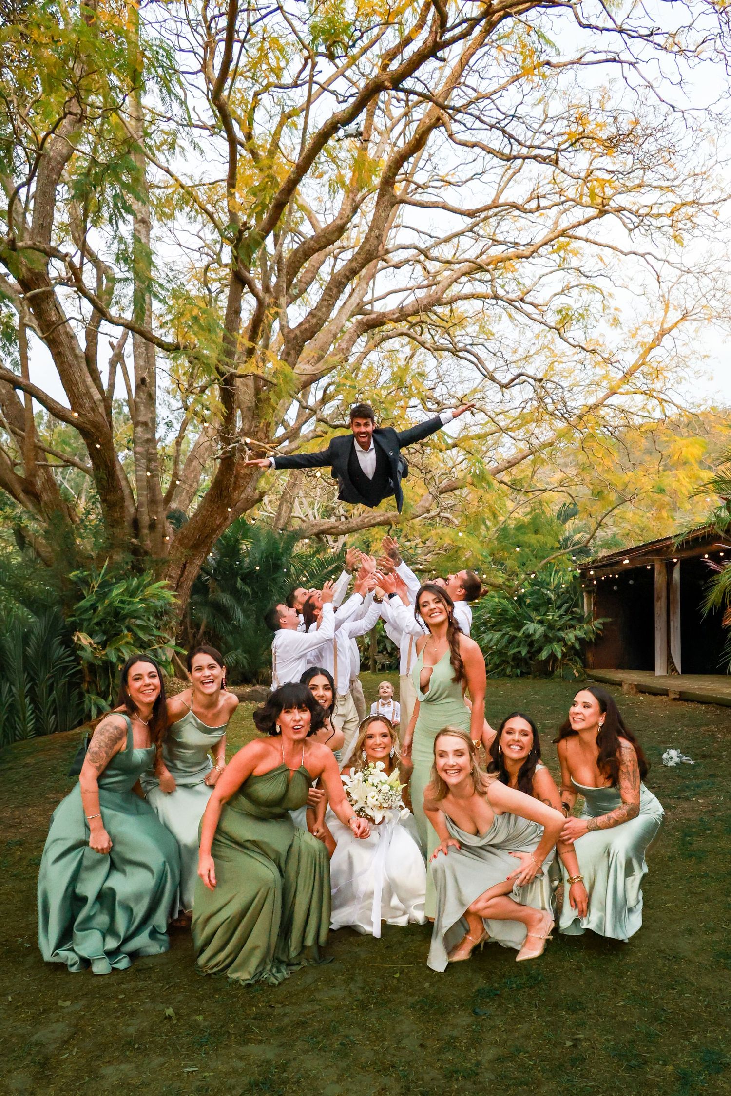 Bridesmaids in sage green dresses pose playfully on grass during autumn wedding photoshoot.