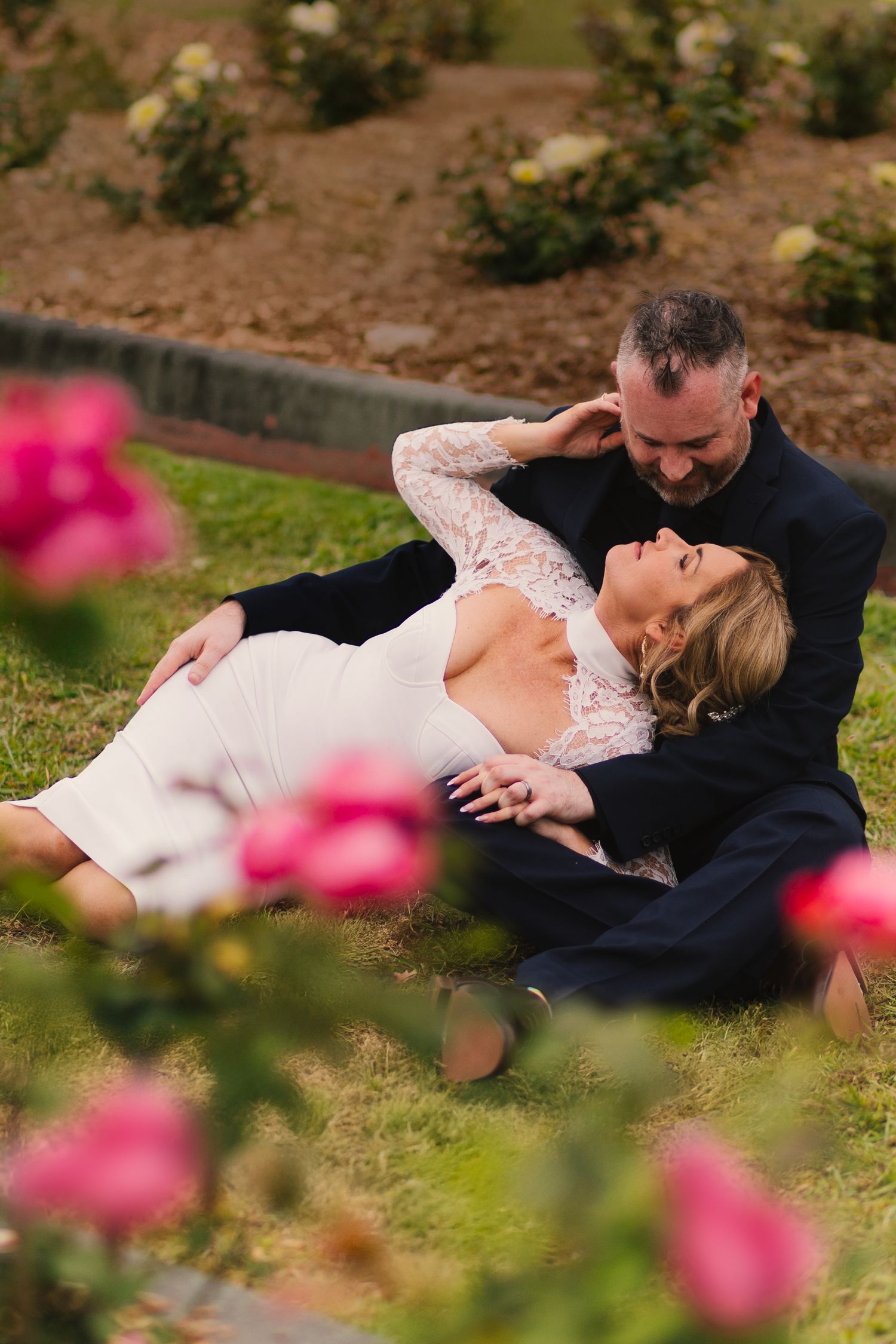 A romantic couple rests together on grass surrounded by pink roses in a garden setting.