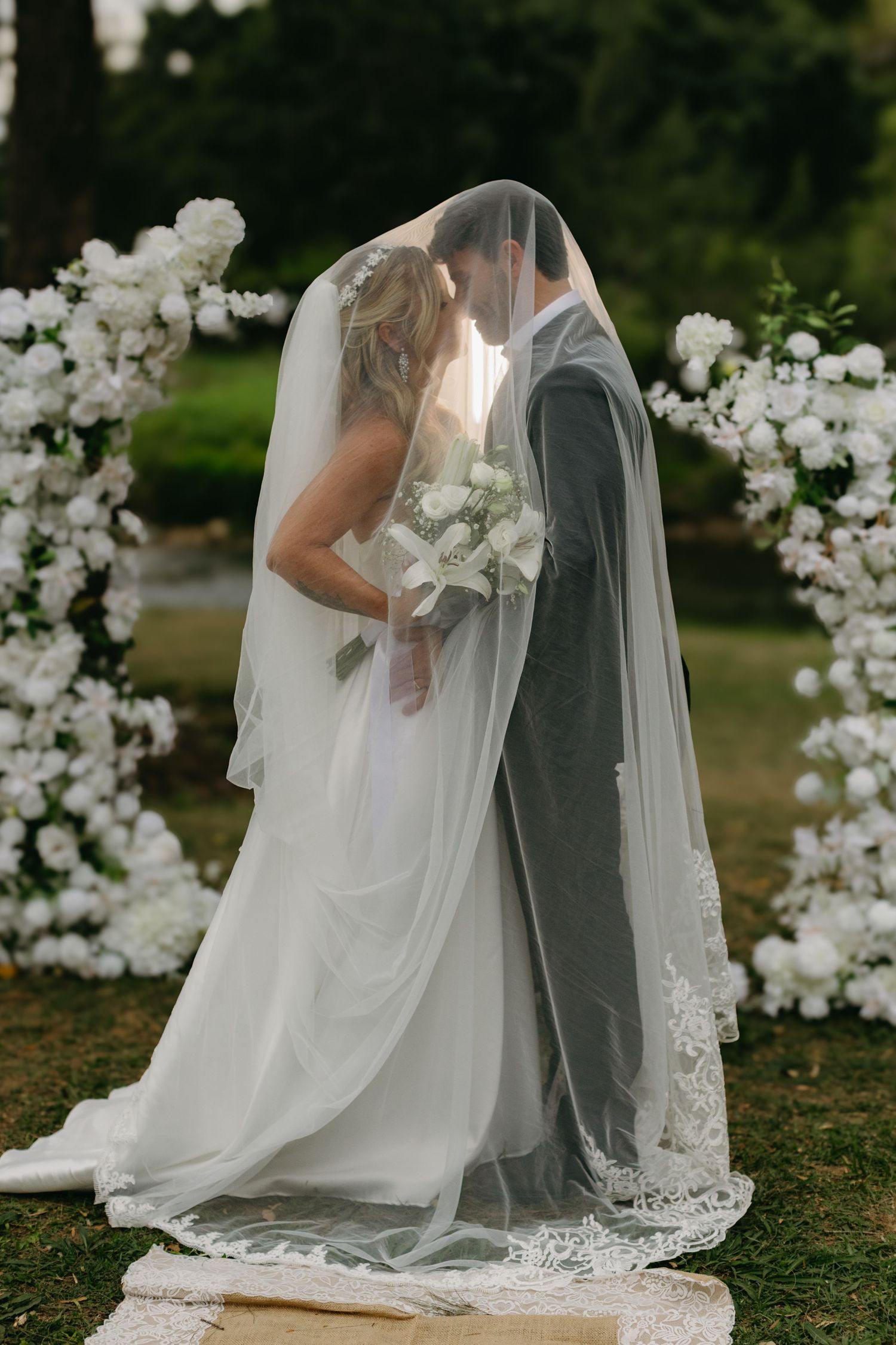 Bride and groom sharing a kiss under a flowing veil at an outdoor wedding ceremony with white floral arch.