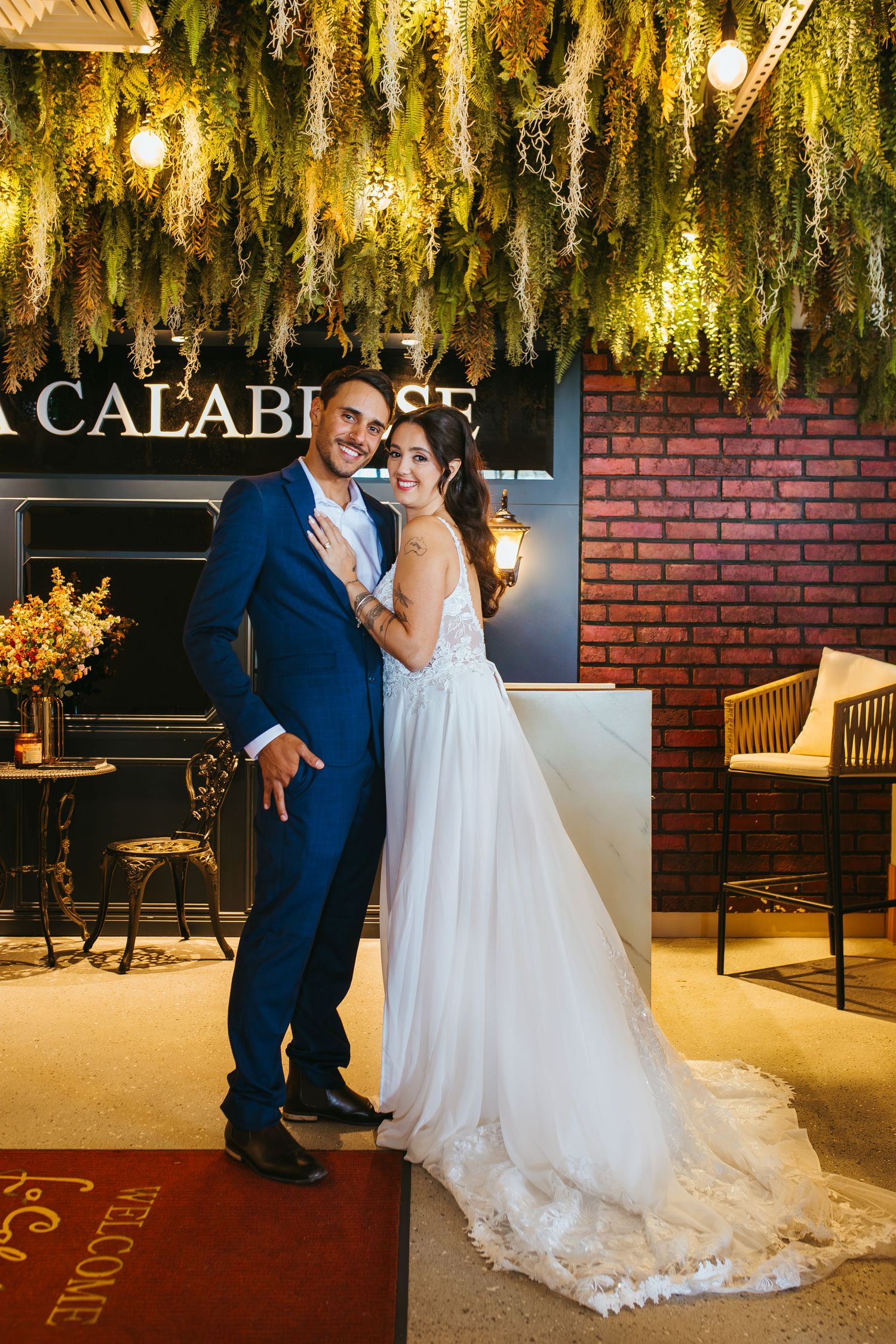 Wedding couple poses against brick wall under hanging golden decorations.
