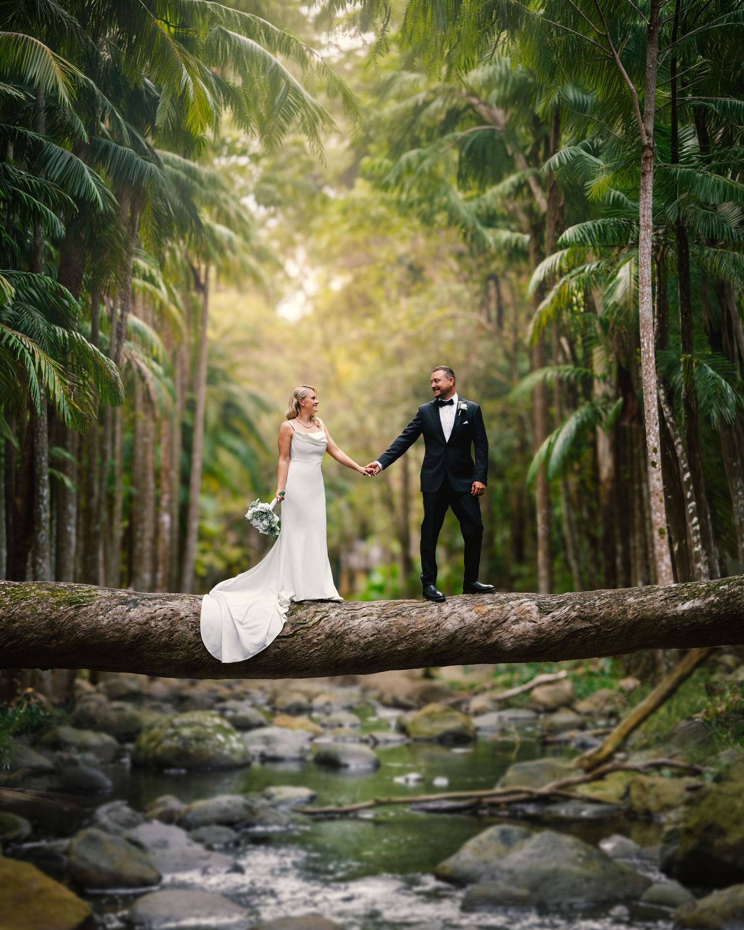 Bride and groom holding hands on a fallen log bridge surrounded by lush tropical rainforest and rocky stream.