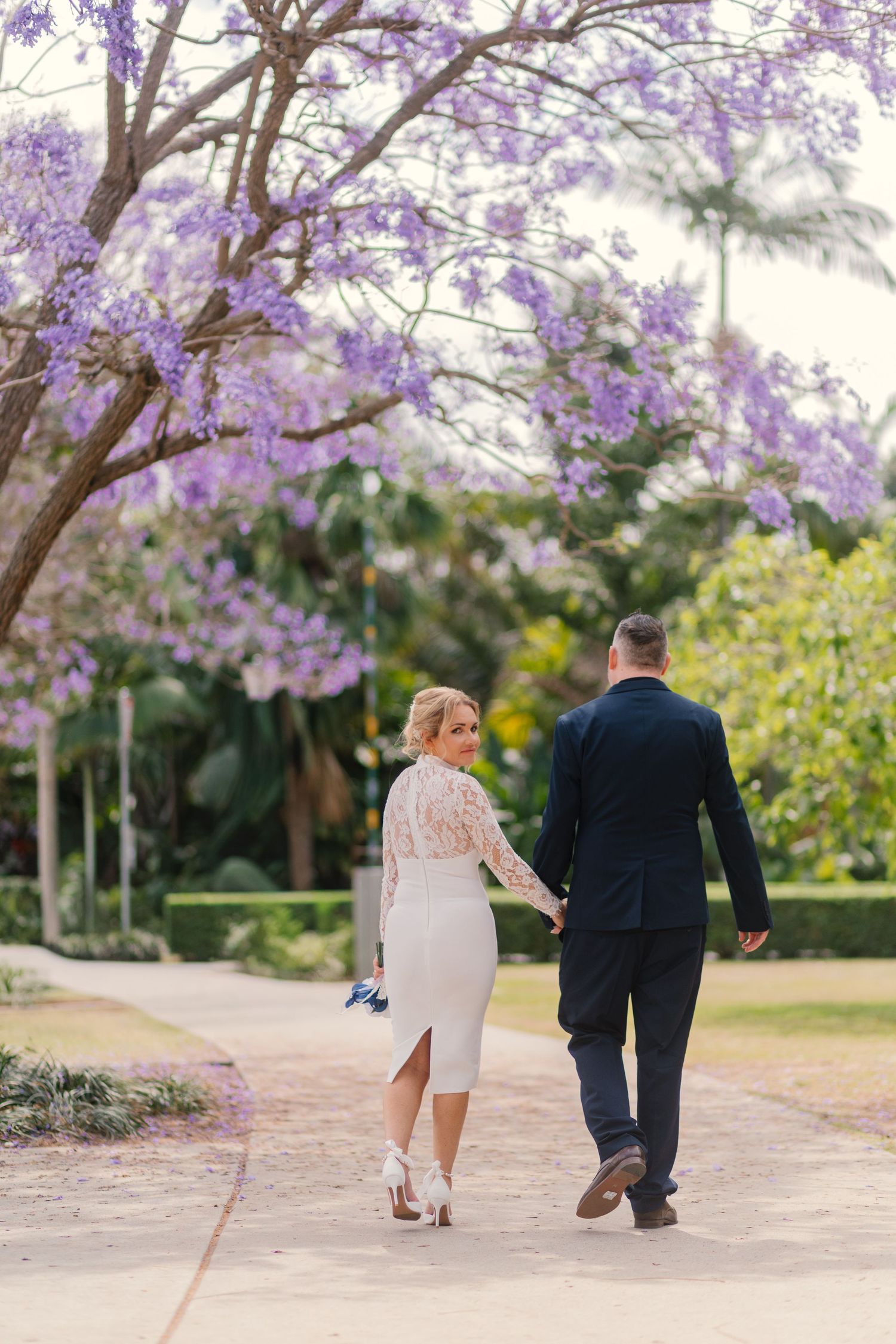 A couple walks hand in hand down a path under blooming purple jacaranda trees in a romantic garden setting.
