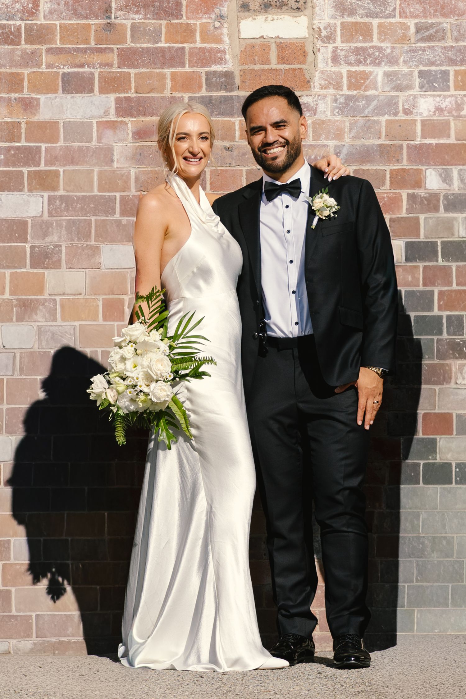 Wedding couple poses against rustic brick wall in formal black and white attire with elegant bouquet.