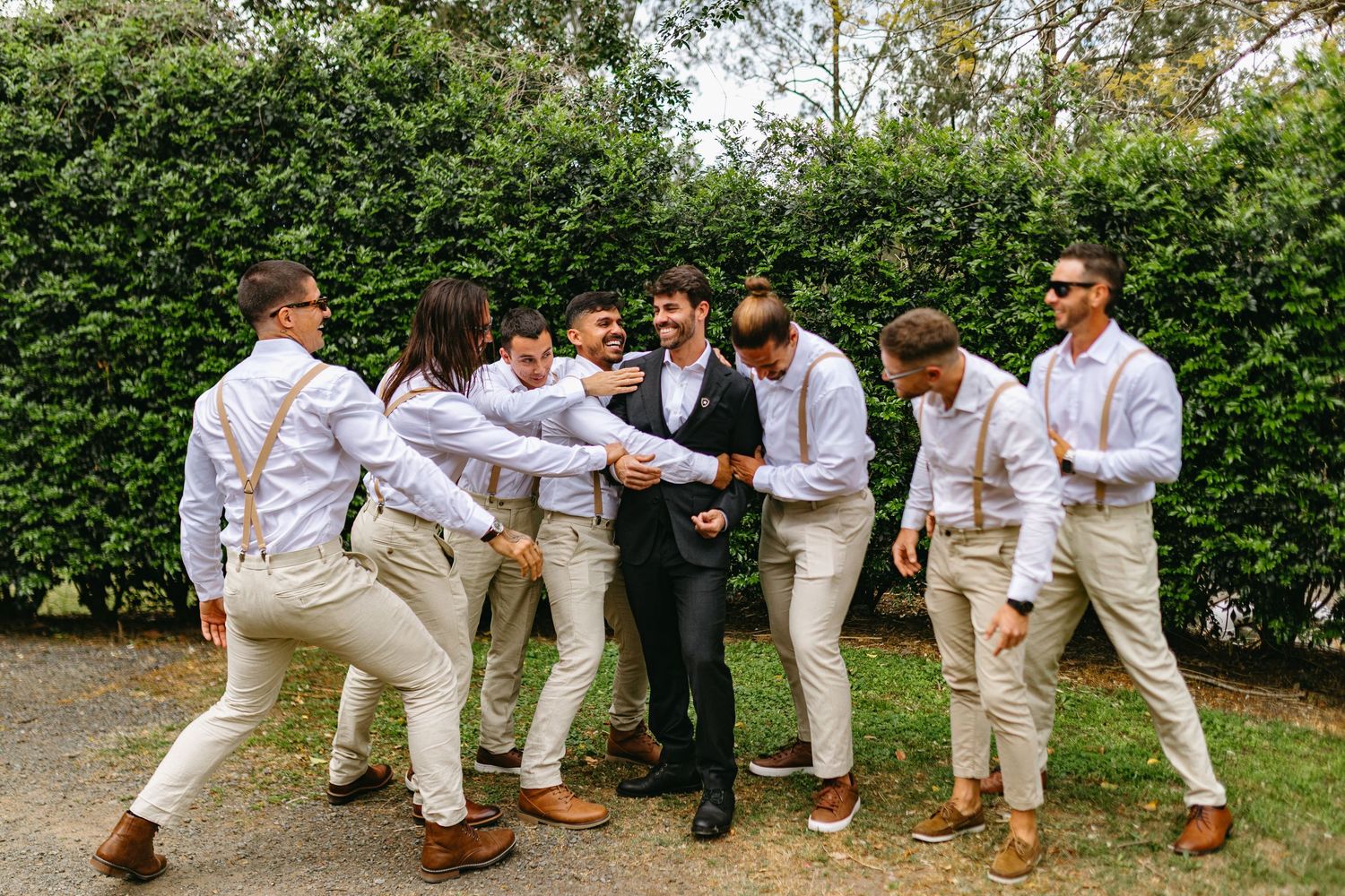 A fun wedding party poses for a series of creative jumping and kicking photos in white and beige attire outdoors.