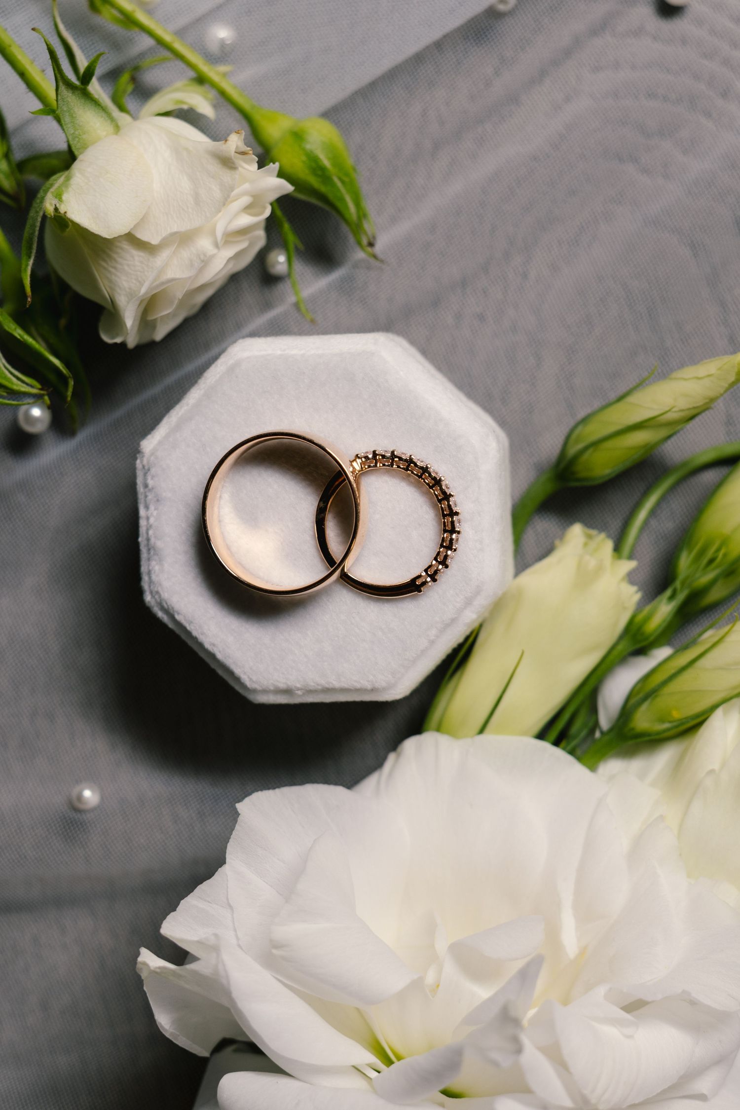 Wedding rings displayed in a white octagonal ring box with white flowers on a gray background.