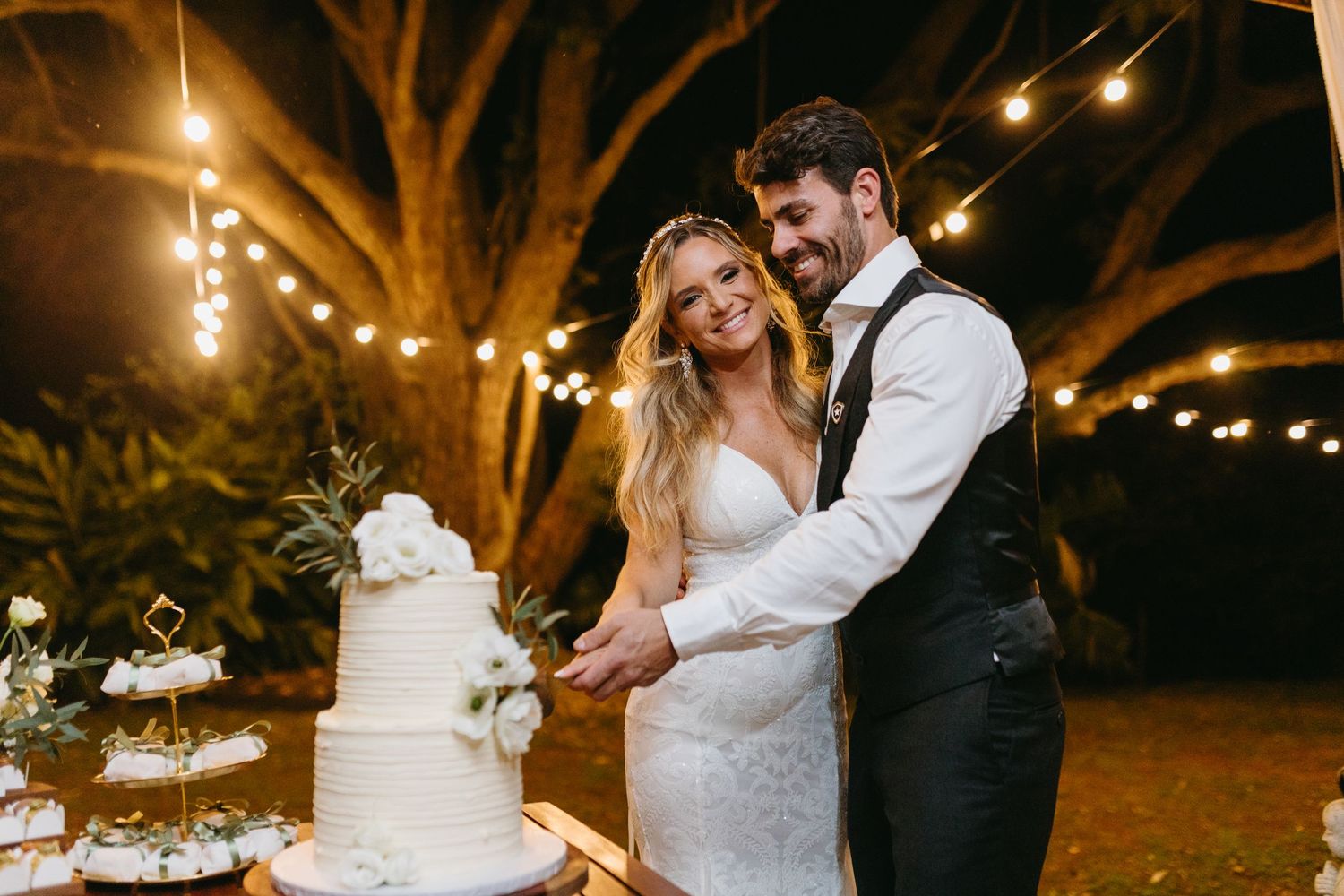 Wedding cake cutting moment under string lights at outdoor evening reception.