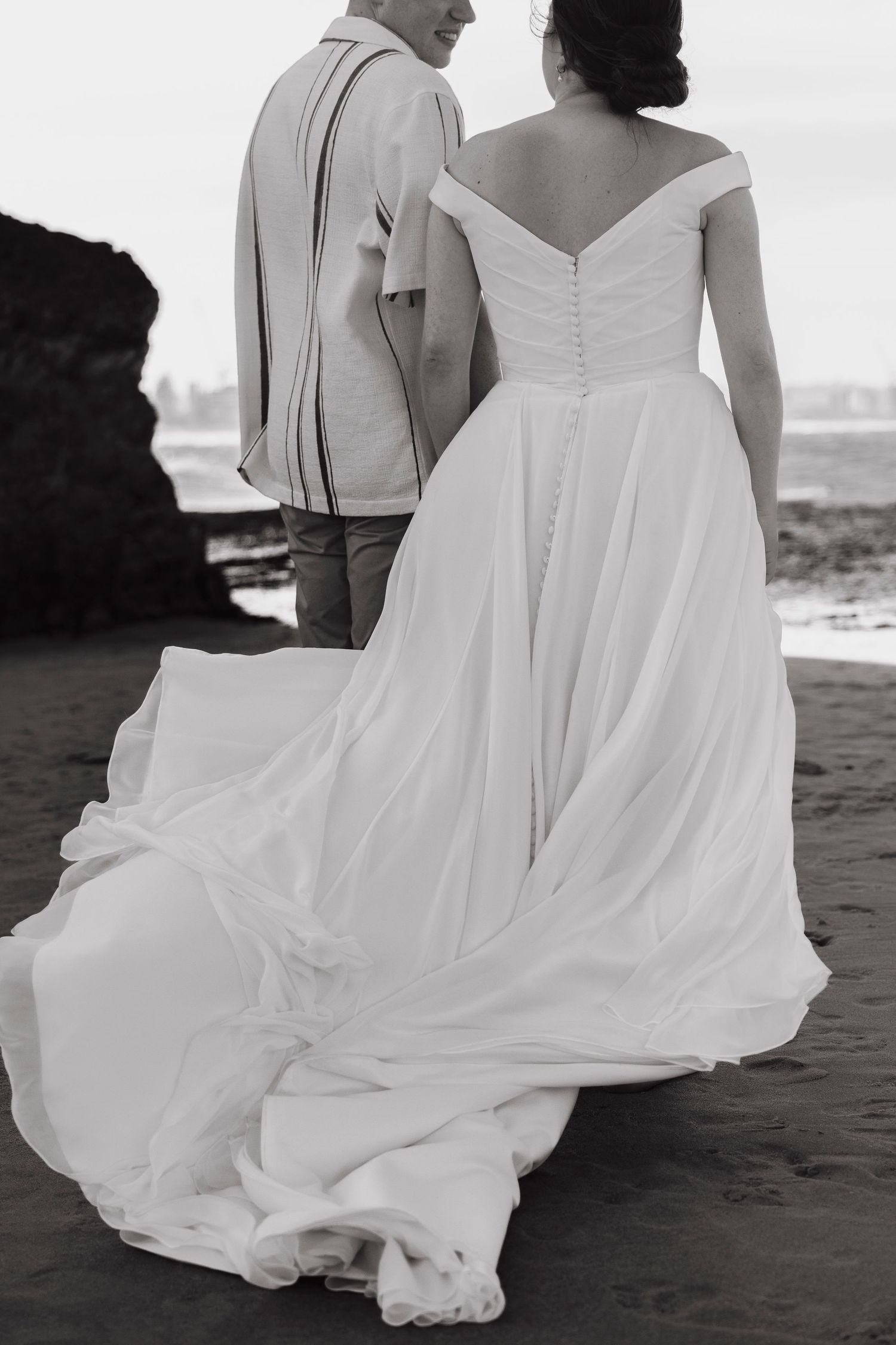 A couple walks along a black sand beach with flowing white wedding dress fabric trailing behind them in black and white.