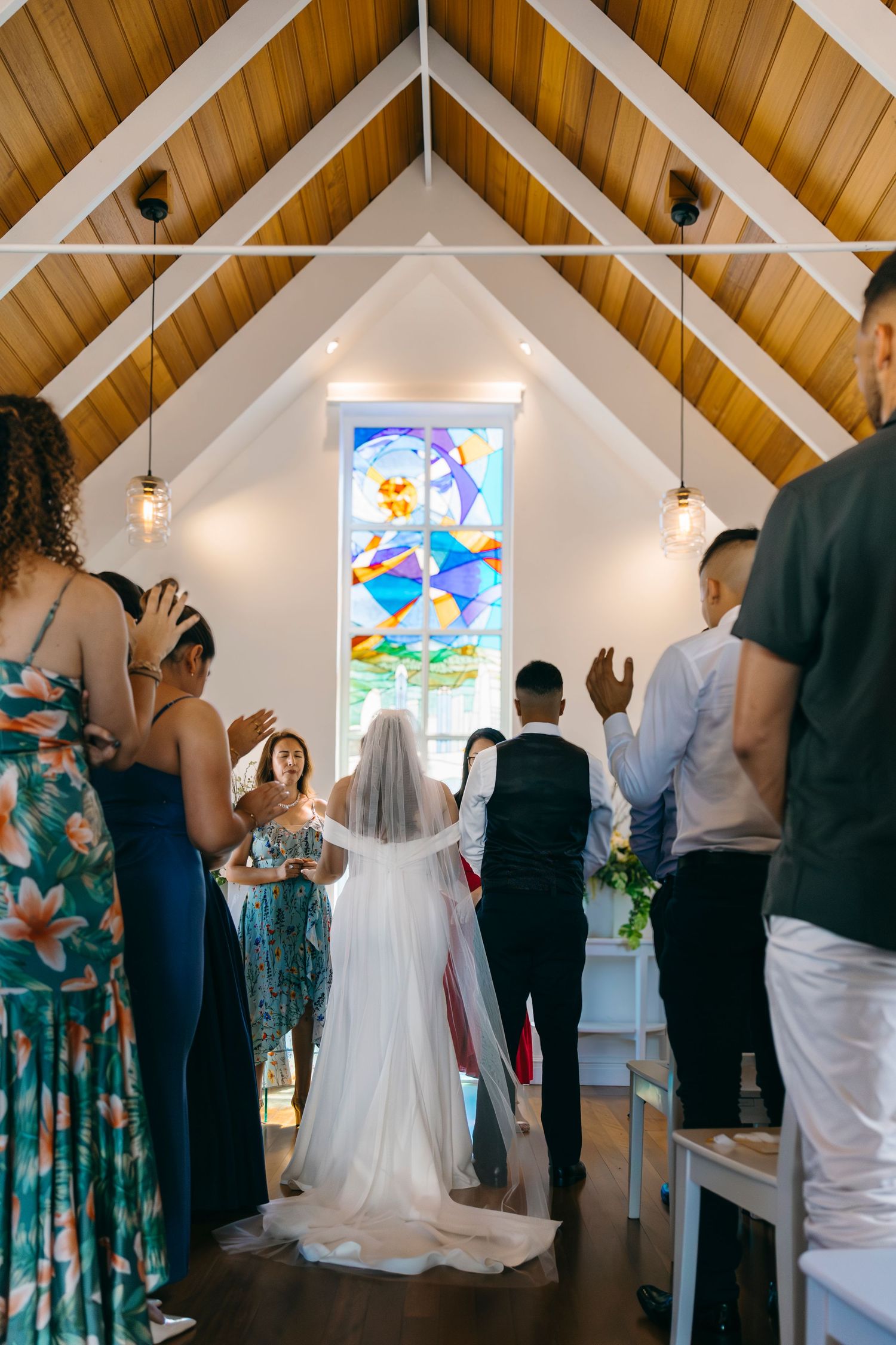 Wedding guests stand in a small chapel with wooden beams and stained glass windows during a ceremony.