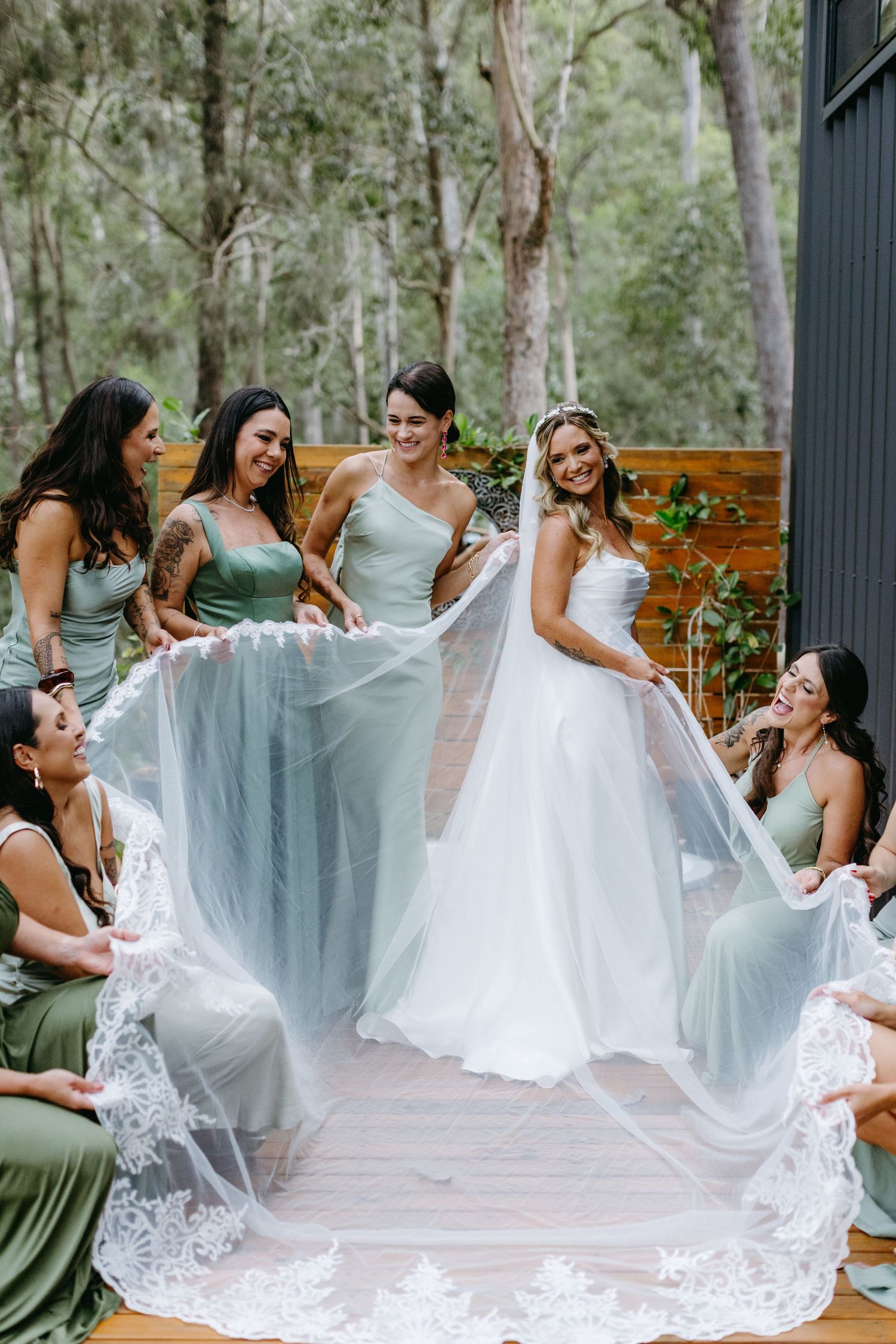 Wedding party members in sage green dresses helping arrange a bride's lace gown.