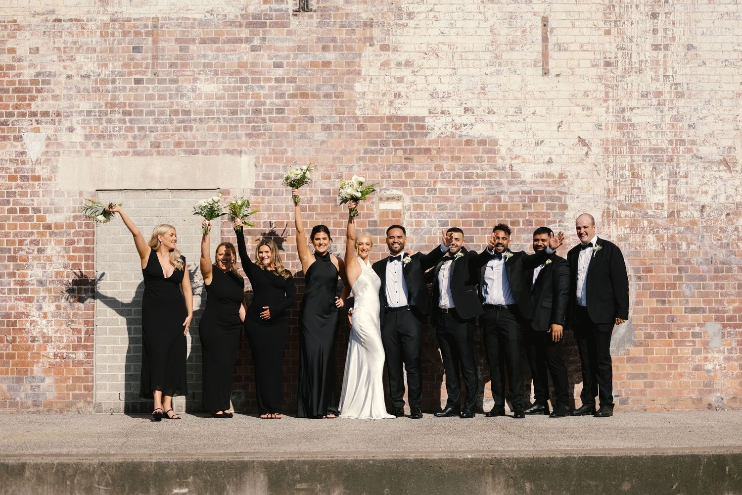 Wedding party poses against rustic brick wall in formal black and white attire.