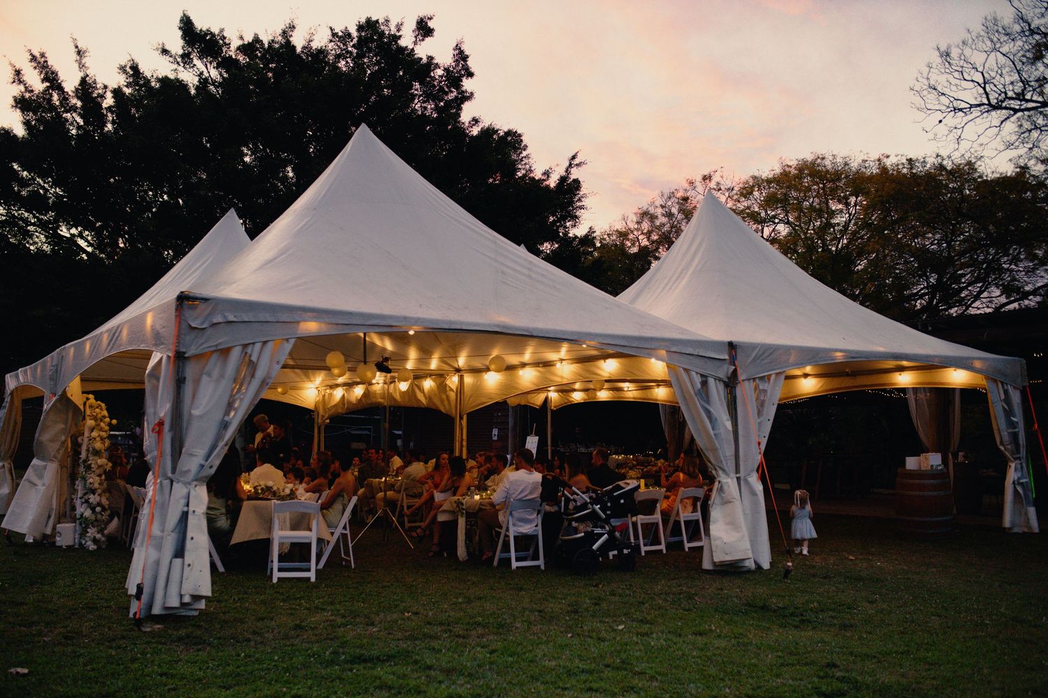 White wedding reception tents illuminated at sunset with string lights creating a romantic outdoor atmosphere.